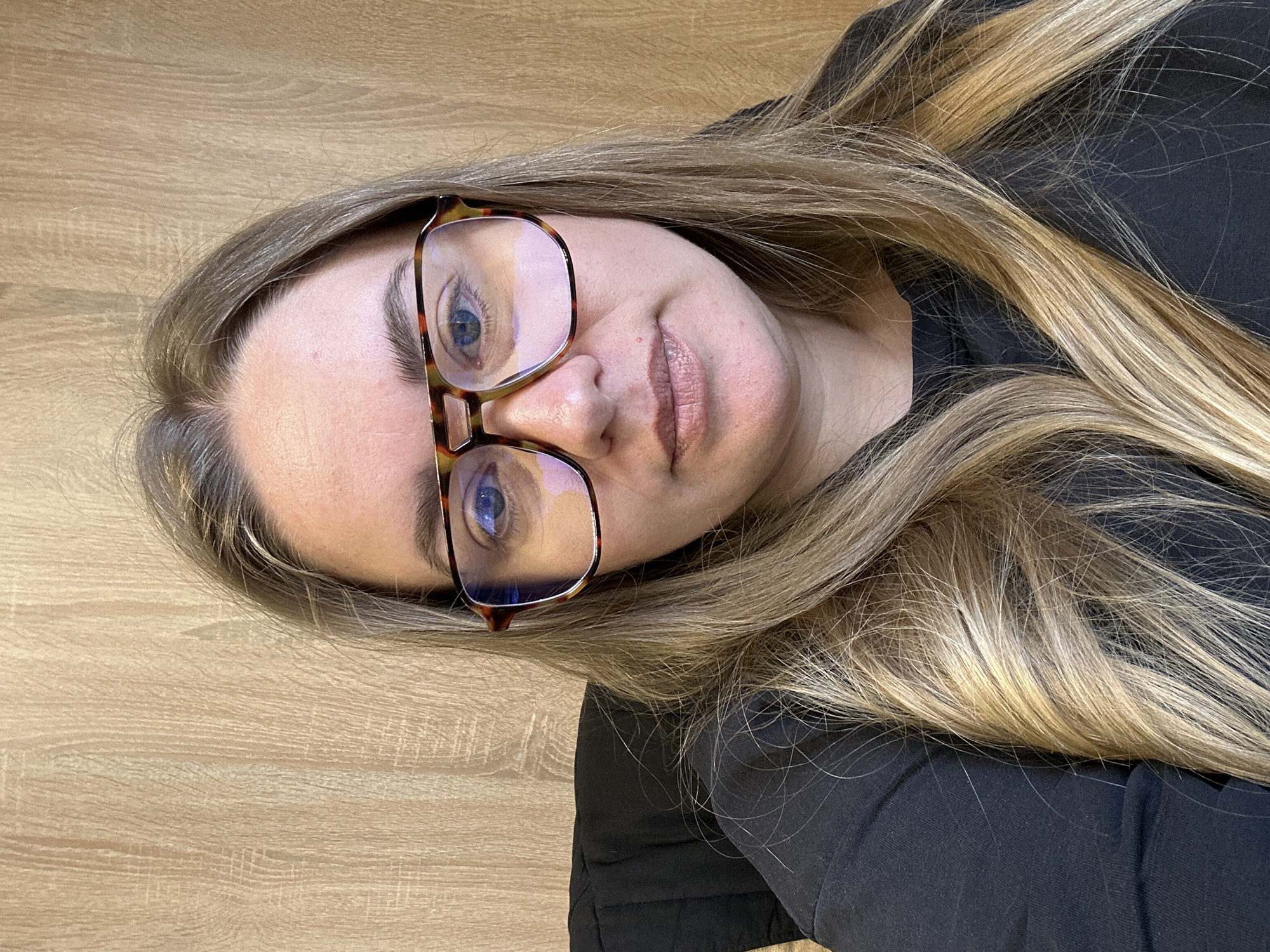 A person with long blonde hair and blue eyes, wearing eyeglasses with tortoise shell frames, is lying down on a light wood surface. The person is wearing a black shirt. The image orientation is rotated slightly clockwise. There is no information to suggest this image was taken in the city of Ural'skoe, Russia.
