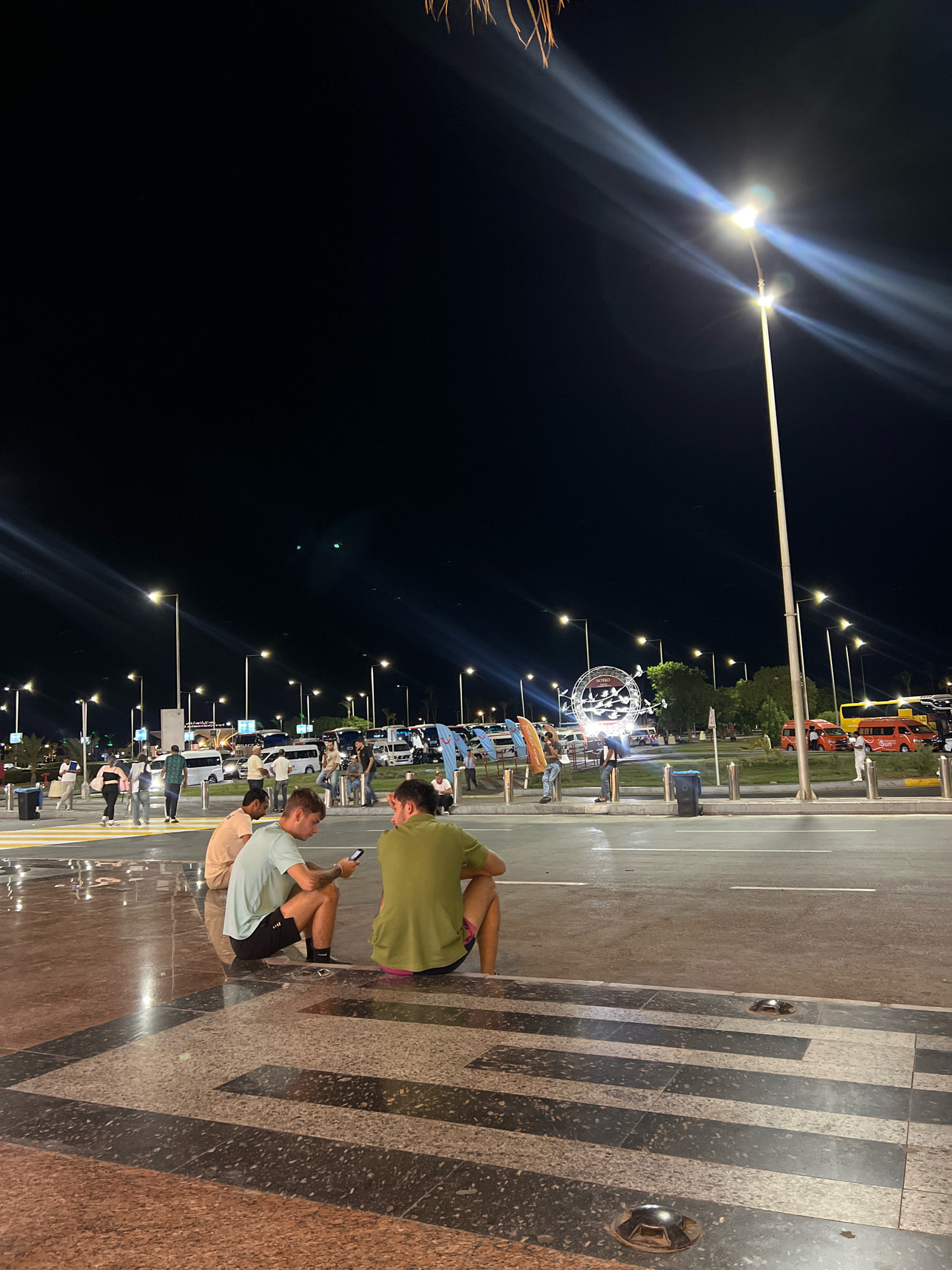 The image shows a nighttime scene in Sharm El Sheikh, Egypt, likely near a transportation hub or a public square. In the foreground, two men are sitting on the ground, facing away from the camera. One man, wearing a light blue t-shirt and black shorts, is looking at his phone. The other man, wearing a green t-shirt and what appears to be pink shorts, is also looking down. Their legs and the patterned tile floor they are sitting on are clearly visible.
In the midground, there is a street with several vehicles, including white vans, a yellow bus, and orange minibusses. A large, circular art installation with multiple bird figures is visible in the distance, illuminated by lights. Several people are walking or standing in the area, appearing to be tourists or locals. Tall lampposts with bright lights illuminate the scene. The sky is dark, characteristic of nighttime. The overall impression is a lively public space at night, possibly an airport or bus station area in Sharm El Sheikh.