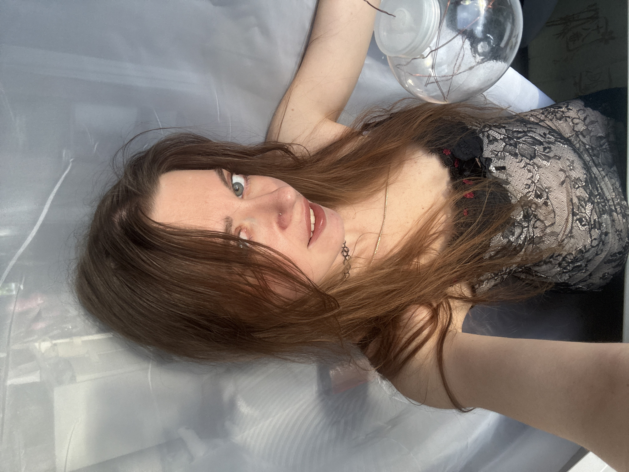 In an indoor setting, a young adult with long, auburn hair and blue eyes is positioned with their body angled towards the camera. They are wearing a black lace corset with floral detailing and a delicate silver choker necklace. Their head is tilted slightly, and they are looking directly at the viewer with a playful expression. A clear glass orb, containing what appear to be copper wires, is held in their right hand. The background is a soft, diffused white, possibly a curtain or sheet, with some shadow play on the left side. The lighting suggests a natural light source, possibly sunlight filtering through a window. The overall scene appears to be a self-portrait.