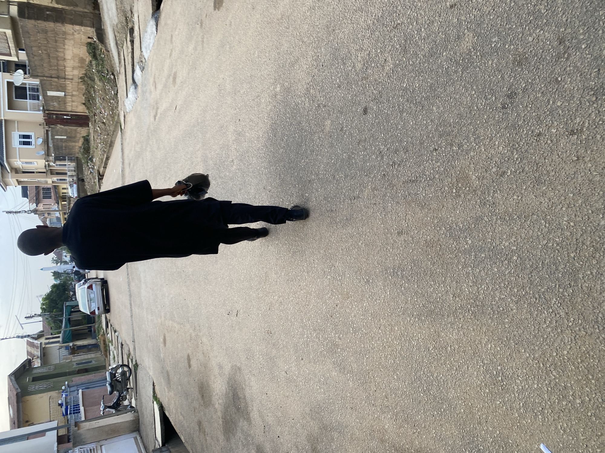 In Jos, Nigeria, a man walks away from the camera on a paved street. He is wearing a black traditional robe and dark pants, and he carries a dark bag in his right hand. The street is lined with houses and a few parked cars, including a white sedan further down the road. A motorcycle is parked on the left side of the street. The sky is overcast.