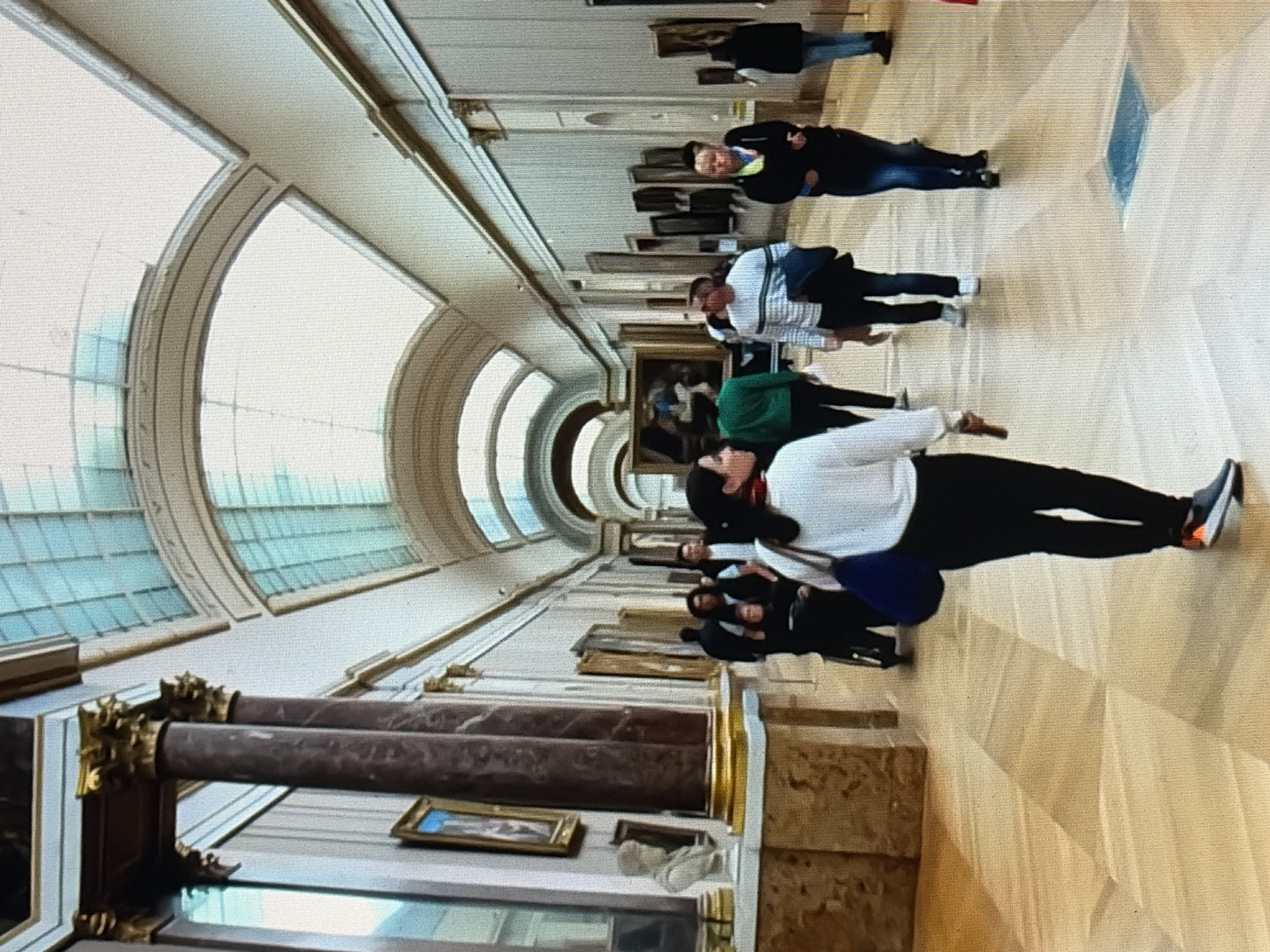 The image shows a wide, well-lit museum hallway with a high, arched, glass ceiling. Several people are walking down the hallway, which is lined with paintings on the walls. The floor is made of polished wood laid in a parquet pattern. In the foreground, a woman in a white sweater and black pants is walking away from the camera. To her left, a large marble column with gold accents is visible. Further down the hall, other individuals are seen browsing the art. A person in a wheelchair is also present. The overall impression is of a busy, elegant art museum.