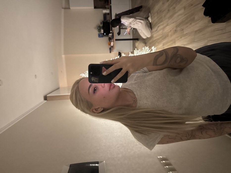 A woman with long blonde hair poses for a selfie, holding a black smartphone. She wears a gray t-shirt, and tattoos are visible on her arm and shoulder. In the background, another person is kneeling near a desk, looking at a device. The room appears to be a modern living space with light-colored walls and wooden floors. A string of white lights is draped across the scene.
