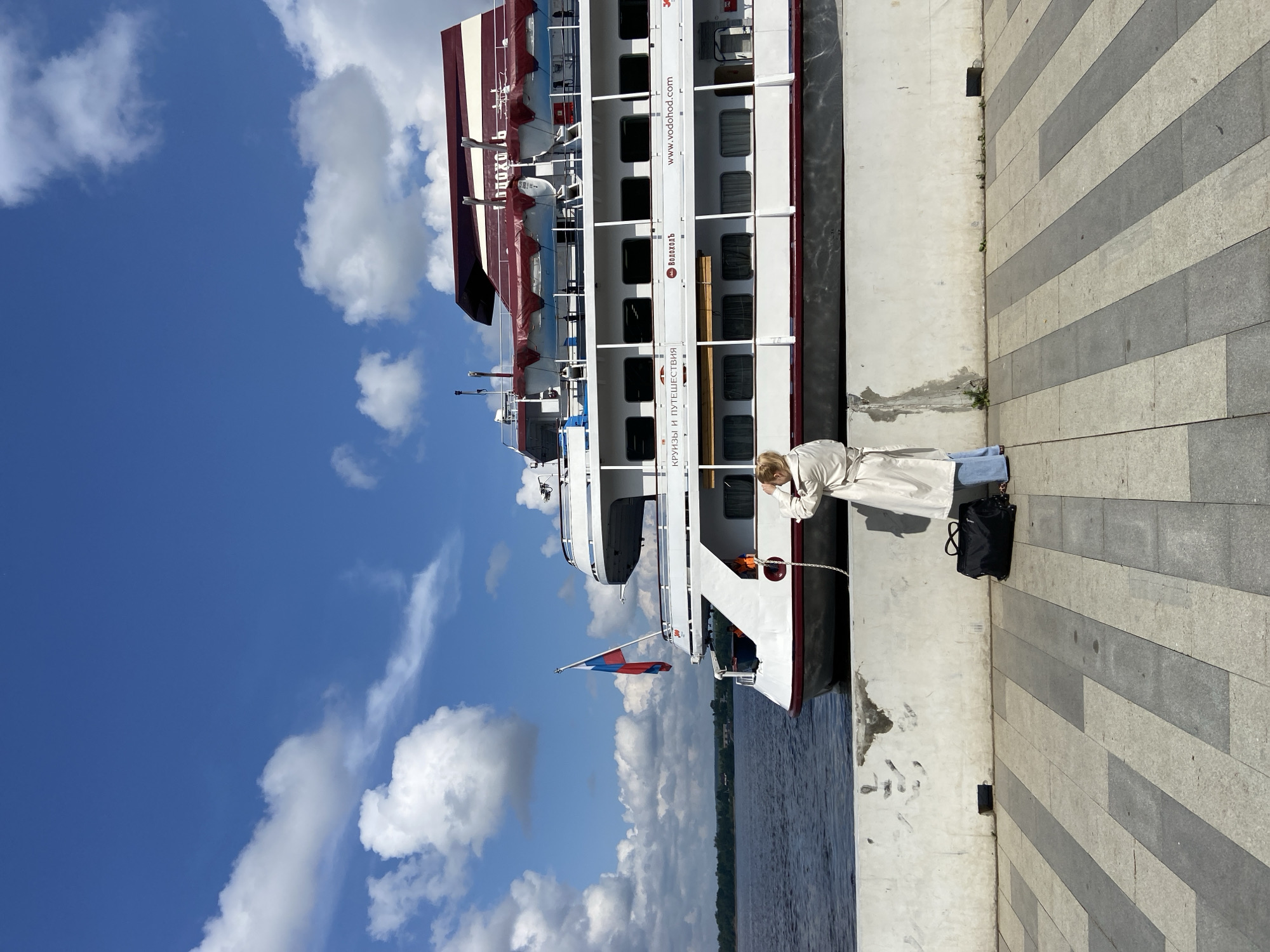 A woman stands on a paved waterfront promenade, facing a large white cruise ship docked on the Kama River. The ship, adorned with a Russian flag and red and white accents, appears to be at a dock in Perm, Russia. The woman, wearing a light-colored trench coat and blue jeans, holds her hand up to her face, possibly adjusting her hair or shielding her eyes. A black duffel bag rests on the ground beside her. The promenade is paved with light and dark gray rectangular stones laid in a striped pattern. Behind the ship, a body of water stretches to a tree-lined distant shore. The sky is a vibrant blue with scattered white clouds. Text on the ship's hull reads "КРУИЗЫ И ПУТЕШЕСТВИЯ" (Cruises and Travels) and "www.vodohod.com".