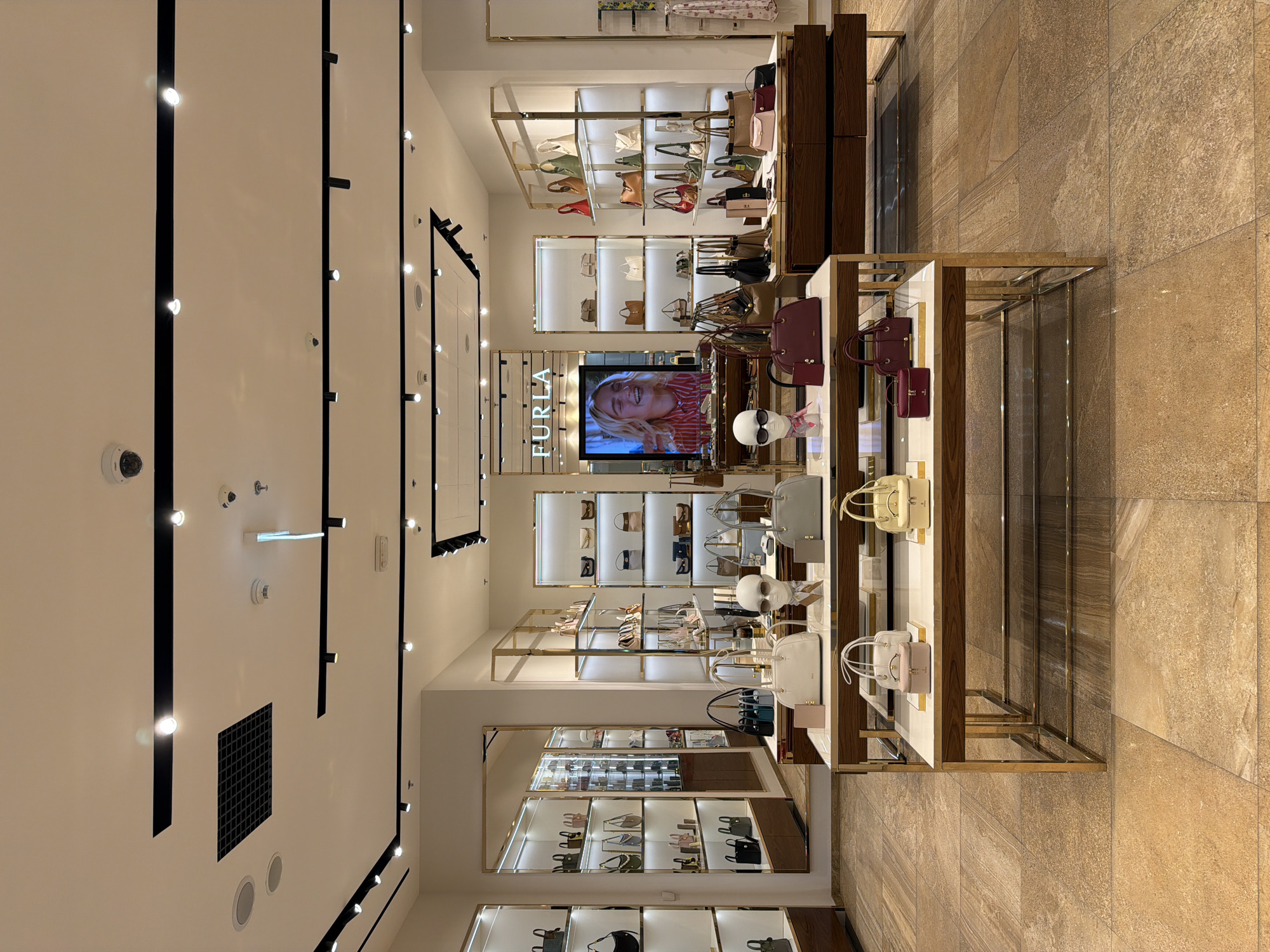 This is a detailed interior view of a Furla retail store in Moscow, Russia, specializing in handbags and accessories. The store presents a modern, upscale aesthetic with meticulous product arrangement and bright illumination.
The foreground features three display tables with white tops, dark wood bases, and gold-toned metal frames. Each table showcases several Furla handbags and small leather goods, including white, yellow, and burgundy pieces, as well as a mannequin head wearing sunglasses and a patterned scarf. The floor is tiled with large, rectangular beige and brown tiles exhibiting a natural stone pattern.
The midground and background reveal numerous display units lining the walls, structured with gold-toned metal frames and illuminated shelving. These units are fully stocked with a diverse array of handbags in various colors, shapes, and sizes. On the left, a large, full-height mirror with a gold frame reflects parts of the store interior. In the center-back, a prominent digital screen displays the "FURLA" logo above an image of a smiling woman, serving as a focal point. Adjacent to the screen are additional shelved displays of handbags and accessories.
The ceiling is white and features a complex lighting system, including linear black track lights with multiple spotlights, circular recessed lights, and dark square ventilation grilles. Several white security cameras and smoke detectors are also visible on the ceiling. The overall lighting is bright and evenly distributed, designed to highlight the merchandise. No individuals are present in the image.