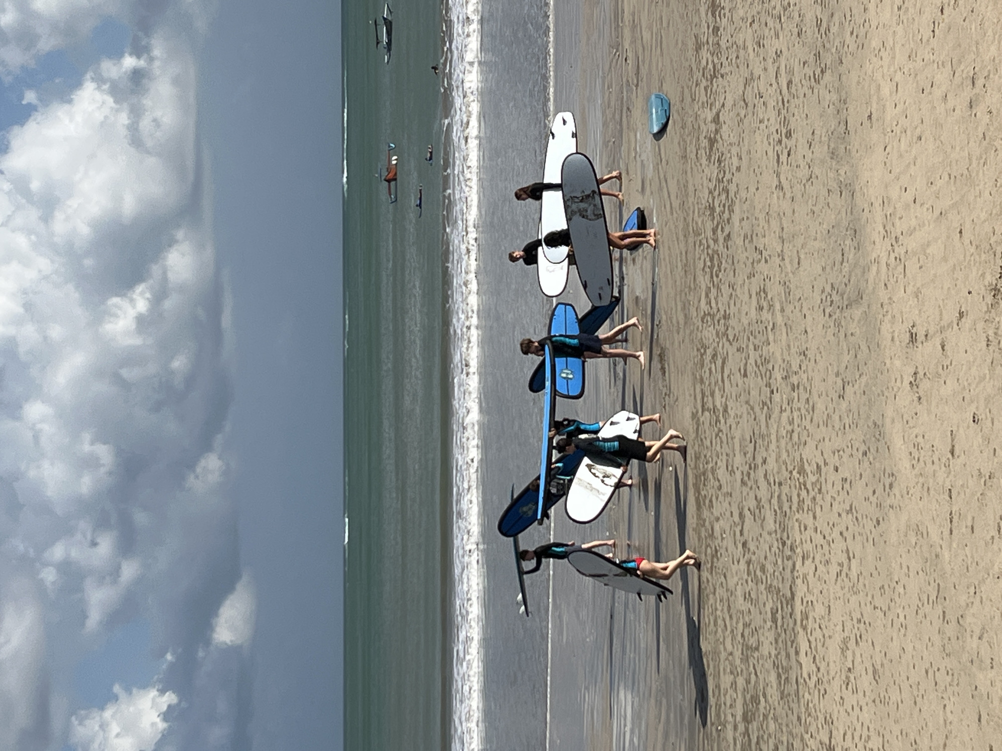 This is a vertical, eye-level, medium shot photograph taken at Kuta Selatan, Indonesia. A group of eight people, appearing to be students and instructors, are walking on a sandy beach carrying surfboards. The water is a deep blue-green, with moderate waves breaking on the shore. The sky is partly cloudy with dramatic, grey clouds dominating the upper portion of the frame. In the distance, several small boats and other surfers can be seen on the water. The overall lighting suggests an overcast day with some diffused sunlight. The sand is damp and reflective in places, indicating the recent presence of water. The surfboards are of various colors and sizes, with several being blue, white, and black. The people are dressed in surf attire, including wetsuits and board shorts. Their movements suggest they are either heading towards or away from the water.