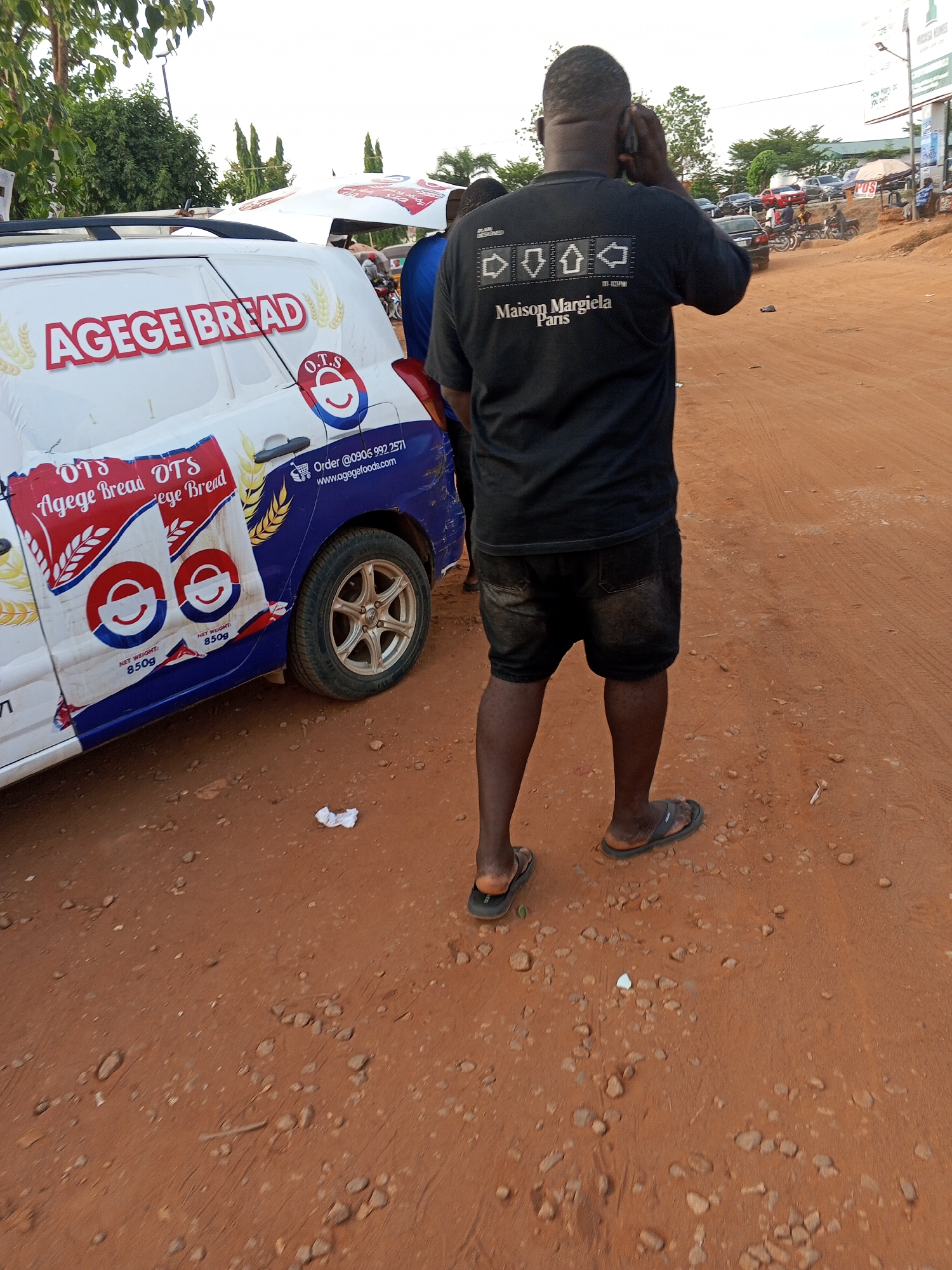 A man in a black Maison Margiela t-shirt and black shorts is on a phone call next to a white van branded "AGEGE BREAD," with an OTS logo, parked on a reddish-brown dirt road. The van displays a phone number and website. The road is uneven, with small rocks and debris. In the background, there are buildings, trees, and other vehicles. A large sign with the text "Micas Homes" is visible. The location appears to be in the city of Galadimawa Village, Nigeria.