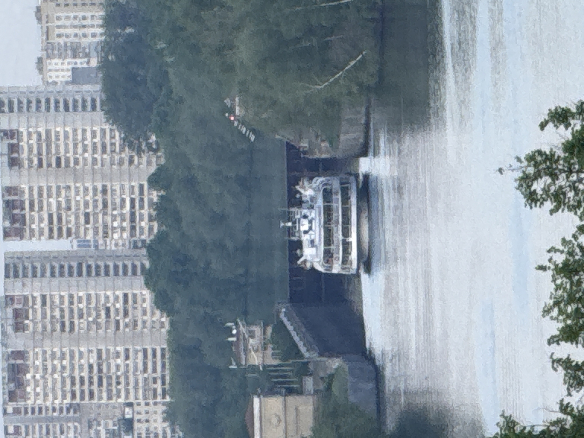 The image shows a cityscape in Moscow, Russia, focusing on a section of the Moskva River. A white riverboat is navigating the waterway, likely a tour or recreational vessel, identifiable by its open deck and visible passengers. On the left side of the river is a modern, high-rise building complex, its facade dominated by numerous windows. The river is flanked by greenery, including trees along the banks, which partially obscure the view. The photograph is taken from an elevated position, offering a view down onto the river and its surroundings.