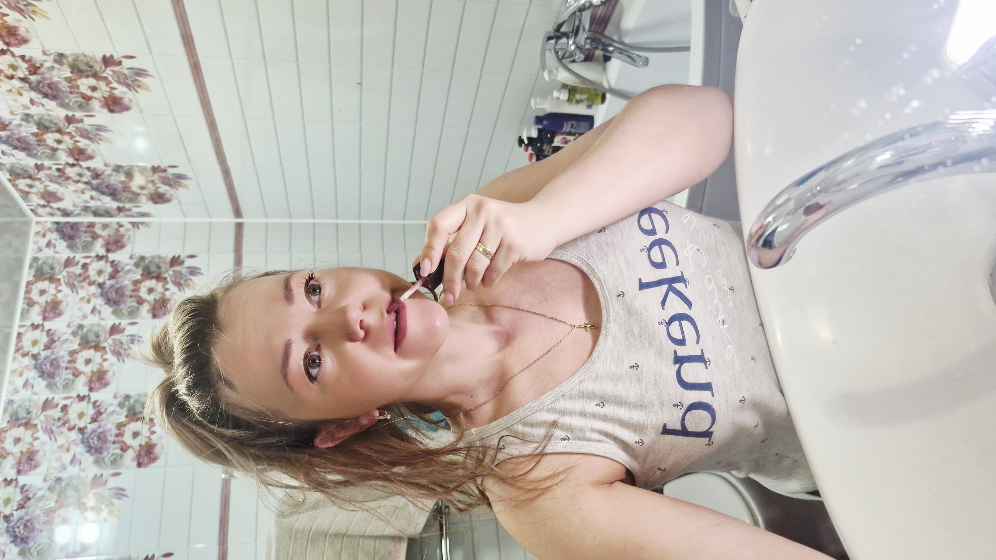 This image, captured in Otchor, Russia, features a young woman in a bathroom setting, engaging in a personal grooming activity. The woman, with light brown hair pulled back and light-colored eyes, is seen applying a light pink lip gloss or lipstick to her lips. She holds the applicator in her right hand, which also bears a gold ring on one finger. A delicate gold chain with a small cross pendant hangs around her neck.
She is wearing a light grey tank top adorned with blue, stylized, and somewhat garbled text, accompanied by small anchor symbols, suggesting a nautical theme. The woman appears calm, looking towards the camera (or into a mirror) with a slight, gentle smile.
The background clearly identifies the setting as a bathroom, dominated by a white ceramic sink and a gleaming chrome faucet. Behind the faucet, several bottles of personal care products are neatly arranged on a shelf. The walls are tiled; one section displays a decorative floral pattern featuring purple, pink, and green hues, while another section consists of plain white tiles accented by a thin reddish-brown border. The lighting is bright and even, indicative of indoor illumination. The photograph itself is oriented 90 degrees clockwise, making the woman appear on her side.