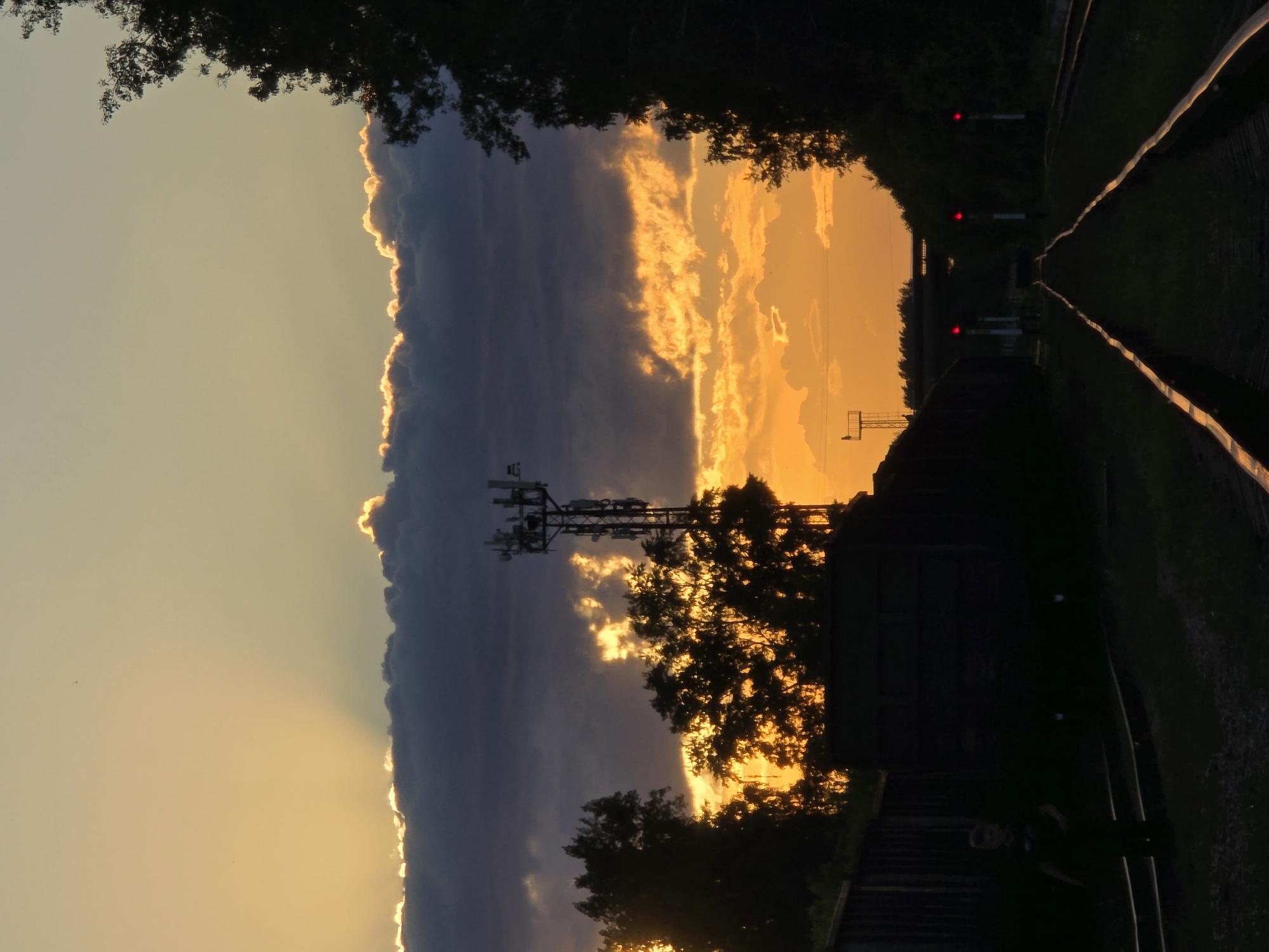 The image captures a dramatic sunset over a landscape in Chaikovsky, Russia, characterized by vibrant colors and strong silhouettes. The sky is the dominant feature, with a large, dark cloud bank stretching horizontally across the mid-section of the frame. Its upper and lower edges are brilliantly illuminated by the low sun, creating a striking golden "silver lining" effect. Above this main cloud, the sky transitions from a soft yellow-orange to a lighter blue, while below it, the sky glows intensely with shades of orange and yellow, featuring scattered, brightly lit smaller clouds.
In the foreground and midground, various elements are rendered in silhouette against the luminous sky. To the left, a tall cell tower, equipped with multiple antennas, stands prominently. Dark tree lines and scattered foliage frame the upper and lower portions of the cloud bank and are visible throughout the midground, especially near the tower. To the right, a railway track recedes into the distance, with its rails faintly reflecting the sunset's golden light. Several red railway signals are discernible along the tracks, suggesting an active line. Indistinct dark structures, possibly buildings or utility boxes, are visible near the tower's base and alongside the railway. In the lower right corner, a faint, silhouetted figure of a person appears to be standing near the railway tracks.
The scene depicts the peaceful close of day, highlighting the beauty of nature's light show juxtaposed with human infrastructure. The overall mood is serene and picturesque. There is no visible text in the image.