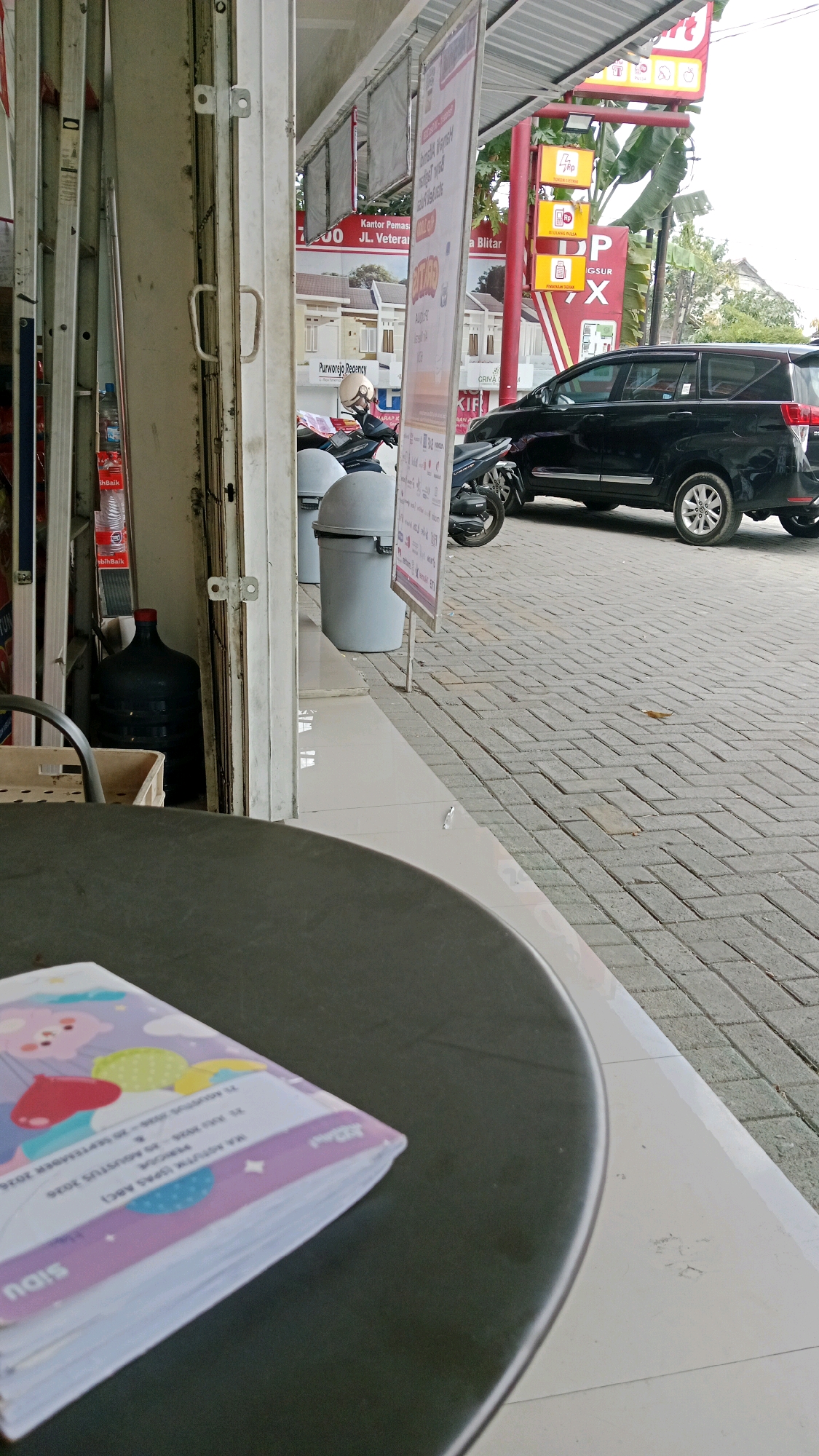 The image depicts an outdoor scene in Blitar, Indonesia, likely in front of a commercial establishment. In the foreground, a dark, round table occupies the lower portion of the frame, with the corner of a book featuring colorful cartoon illustrations and text visible on its surface. The book appears to be a notebook or a similar item with pages for writing.
Behind the table, a white tiled walkway leads to the entrance of a building, partially obscured by a large, black water jug and a metal shopping basket. To the left, a silver ladder is propped against the building's facade. Further back, several light gray trash cans are positioned near the entrance.
In the mid-ground and background, a black SUV is parked on a paved area constructed with interlocking bricks. A motorcycle is parked next to the SUV. The building itself features a striped awning with advertising banners attached. One banner, in particular, displays text in Indonesian, with the word "Blitar" clearly visible, indicating the city of its location. The environment suggests a commercial or retail setting, possibly a convenience store or a similar business, given the presence of goods, advertising, and parked vehicles. The sky is overcast, suggesting a cloudy day.