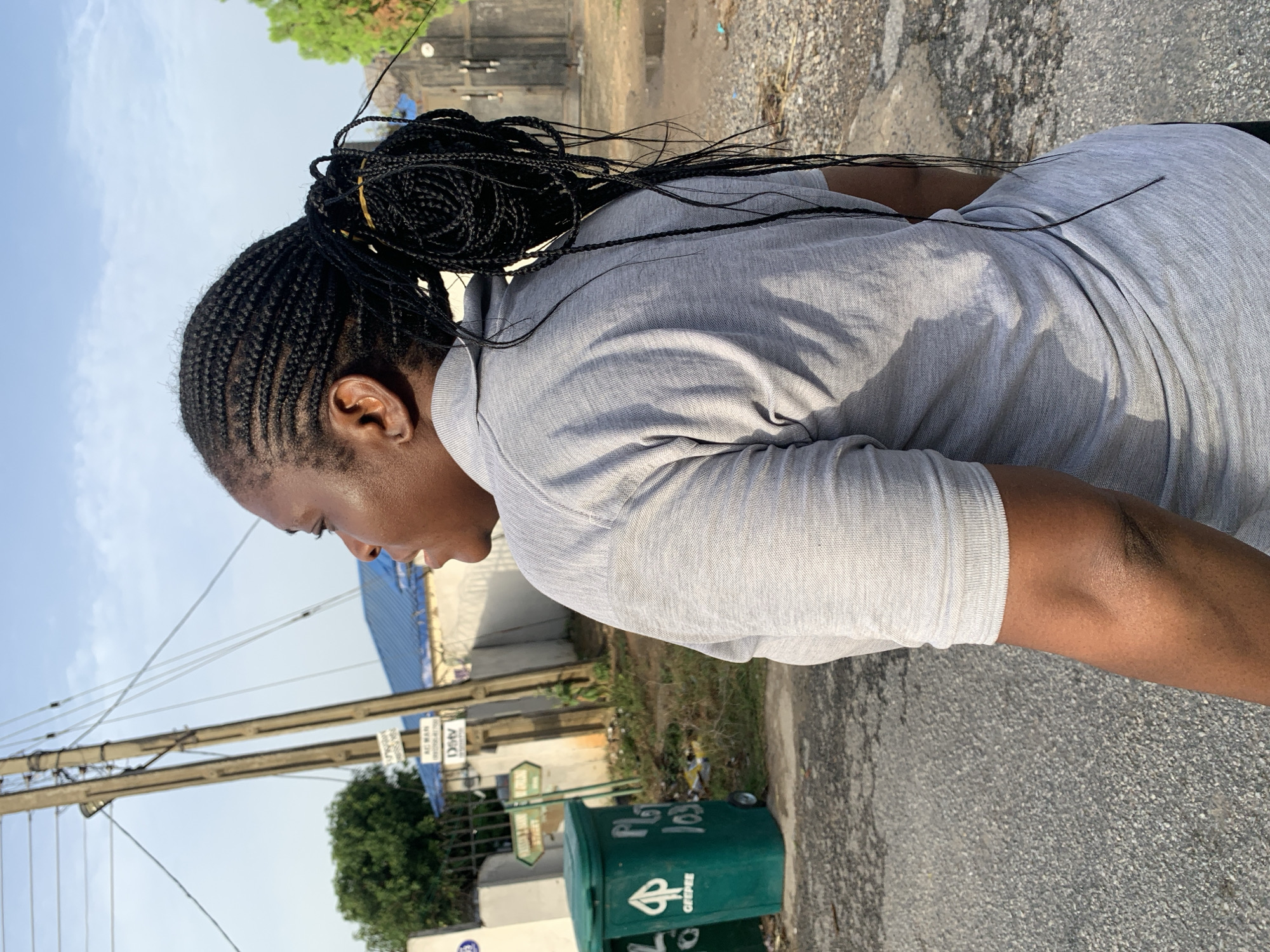 A medium shot shows a woman standing on the side of a road. She is facing left, with her head bent slightly down. Her hair is braided and pulled back into a bun. She is wearing a grey t-shirt.
The background includes power lines, a green trash can, and a building with a blue roof. The setting suggests a location in Abuja, Nigeria, given the architectural style and vegetation common in the city.