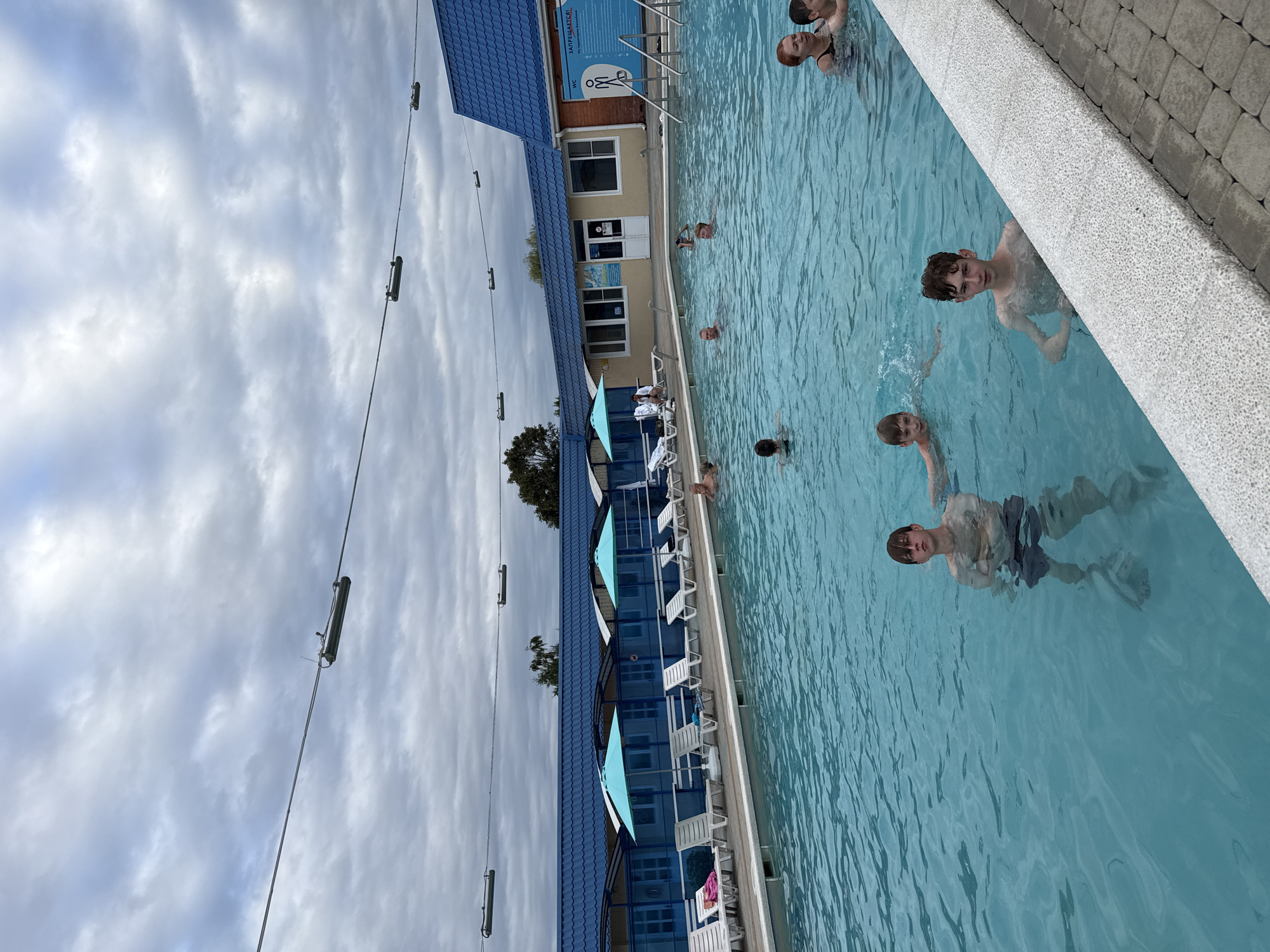 In an outdoor swimming pool facility, several individuals are seen enjoying the water. The pool itself is large and filled with clear blue water. Several lounge chairs are arranged along the perimeter of the pool, some with umbrellas providing shade. In the background, a building with a blue roof and beige walls is visible, which appears to house changing rooms or other amenities. There are also signs posted on the building, one of which indicates "WC" and another with a prohibition symbol. The sky above is cloudy, suggesting overcast weather. People are scattered throughout the pool, some swimming, others standing in the water. The scene is set on a warm day, and the overall impression is one of a recreational area being utilized by its visitors.