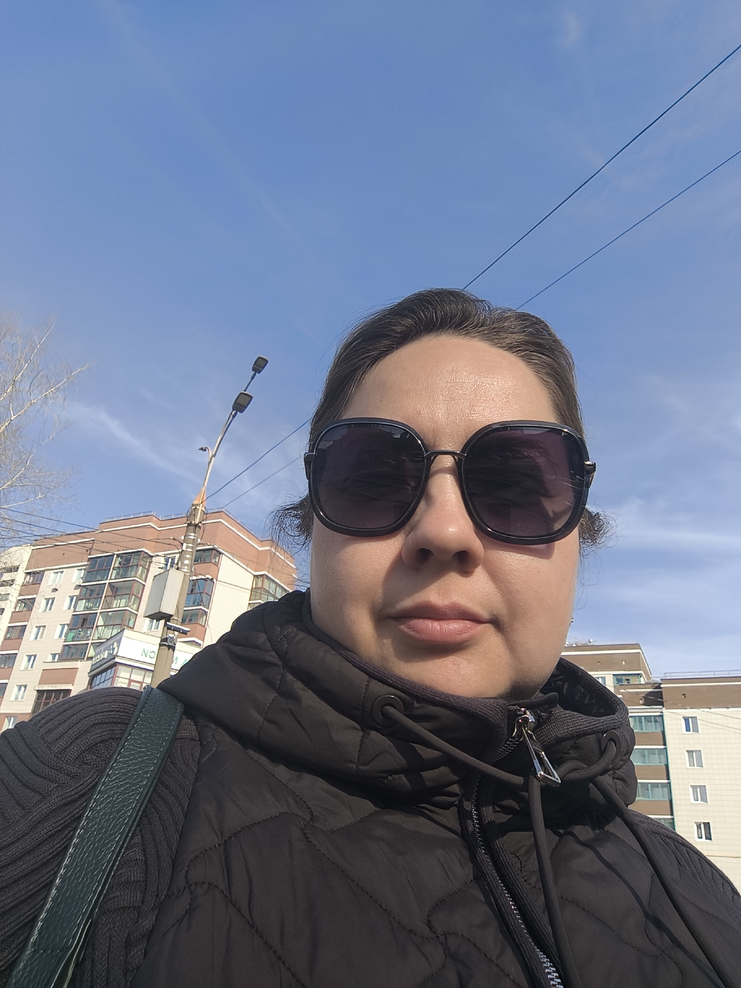 An adult female is depicted in a close-up, selfie-style photograph taken outdoors in an urban setting within Izhevsk, Russia, under a clear blue sky. The individual wears dark-tinted, large-framed sunglasses that obscure her eyes, a black quilted jacket, and a dark, possibly ribbed, garment underneath. A dark shoulder strap, consistent with a bag, crosses her left shoulder. Her dark hair is pulled back, with some wisps visible near the ears. In the background, to the left, is a multi-story residential building with a reddish-brown upper section and lighter lower floors, featuring numerous windows and balconies. A sign with partially visible Cyrillic text (starting "NO M") is present on the lower part of this building. A tall streetlamp pole with two fixtures extends upwards behind the individual. To the far left, a bare-branched tree is visible. In the upper right background, another multi-story building with light-colored facades and windows is partially visible. Two thin, dark lines, likely utility wires, traverse the upper sky. The scene is illuminated by bright daylight, and the sky is predominantly clear with minimal scattered clouds.