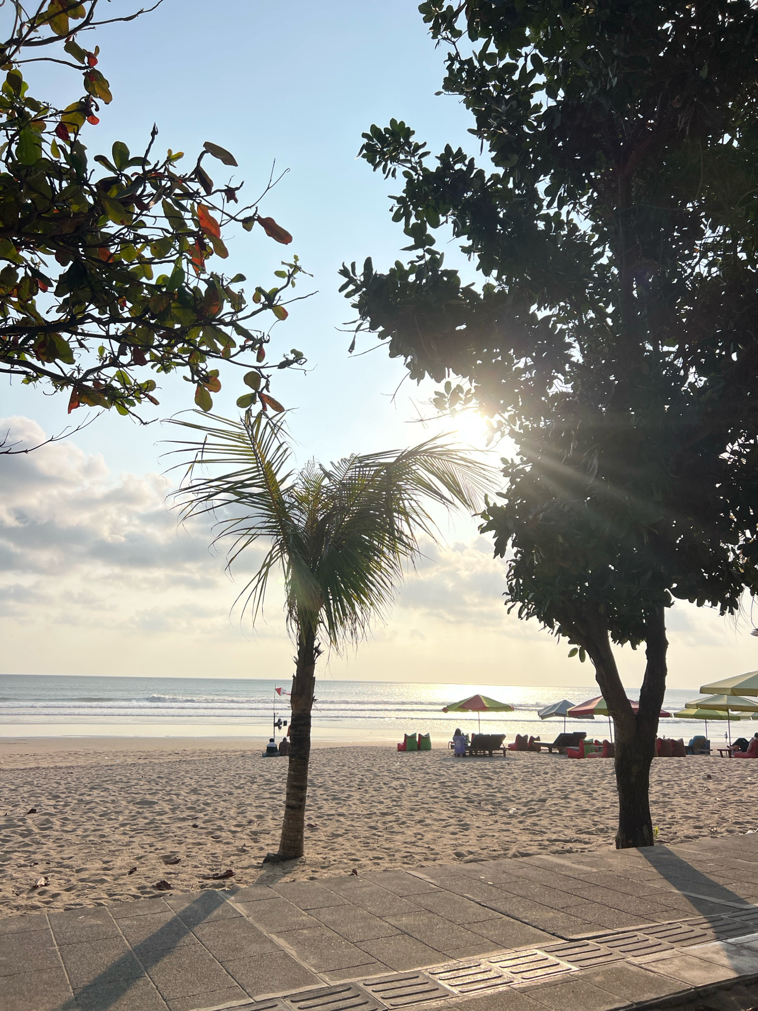 The image depicts a serene beach scene in Kuta, Indonesia, bathed in the warm glow of the late afternoon sun. The foreground features a paved walkway with visible shadow patterns from the surrounding trees. Beyond the walkway, a sandy beach stretches towards the ocean. A lone palm tree stands tall on the sand, its fronds reaching towards the sky. In the mid-ground, beach chairs and colorful umbrellas are arranged, suggesting a popular tourist spot. A few people are scattered across the beach, some resting on lounge chairs, while others are gathered near the water. The ocean is calm, with gentle waves rolling towards the shore. The sky is a soft blue, with scattered clouds catching the sunlight. The overall atmosphere is peaceful and inviting, capturing the quintessential beauty of a tropical beach in Kuta.