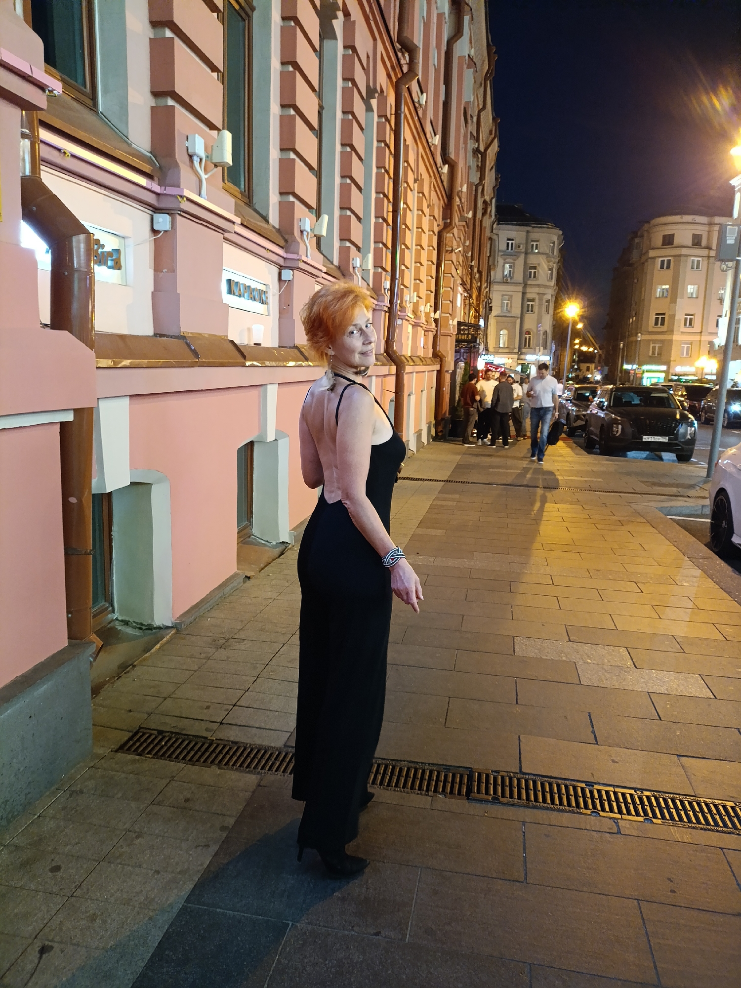 The image depicts an urban street scene at night in Moscow, Russia. The primary subject is a woman with reddish-orange hair and fair skin, positioned on the left side of the sidewalk, looking back over her right shoulder towards the camera with a slight smile. She is wearing a sleeveless, backless black gown and black high-heeled shoes, with a dark choker necklace and a silver-toned bracelet on her left wrist.
To her left, a multi-story building with a light pink facade features decorative, darker pink rectangular pilasters and white trim. Multiple windows with dark frames are visible, along with a metallic downspout running vertically. Signs on this building include "KARAOKE" and a partial sign that appears to be "Бара" (Bar). Small, light-colored electronic devices or sensors are mounted on the facade.
The sidewalk in the foreground is paved with rectangular grey and brown tiles, interrupted by a long, narrow metal drain grate. Warm artificial light from streetlights and building interiors illuminates the scene, casting shadows on the ground.
Further down the street, several men are visible. One man in a white shirt and jeans walks towards the camera, carrying a dark bag. A group of other men stands near an entrance on the left side of the street.
On the street to the right, a dark-colored Hyundai Palisade SUV is visible, bearing a Russian license plate starting with "Y710" and ending with "777RUS", which is characteristic of the Moscow region. A light-colored car is partially visible on the far right. Across the street, other multi-story buildings with varied architectural styles are present, including one with a distinct rounded corner. The sky above is dark, indicating late evening or nighttime.