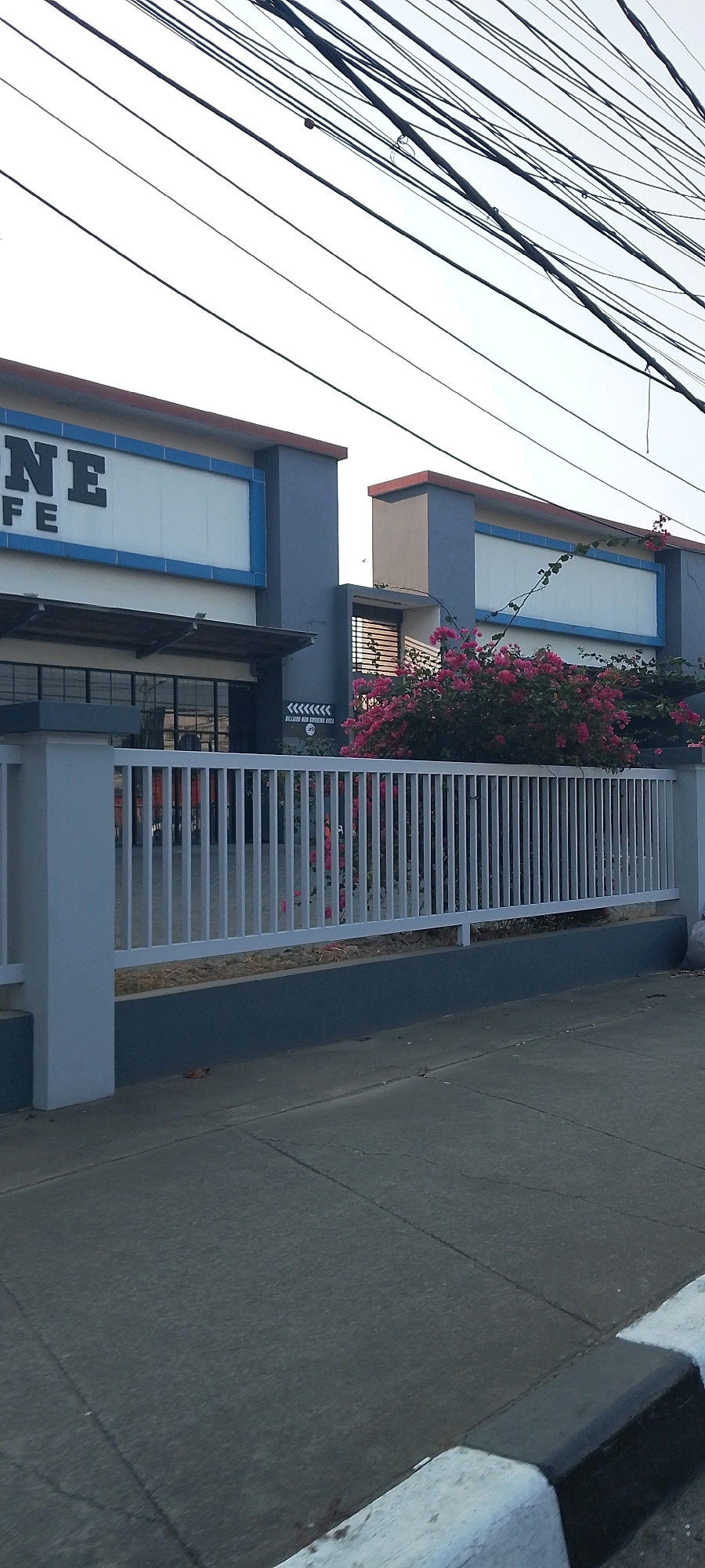 The image shows the exterior of a building with a sign that reads "ONE CAFE". The building is located in the city of Bekasi, Indonesia. There is a white metal fence in front of the building, and behind the fence, there are pink bougainvillea flowers. The sky is visible in the background, with a multitude of electrical wires crisscrossing overhead. The foreground shows a concrete sidewalk with a curb.