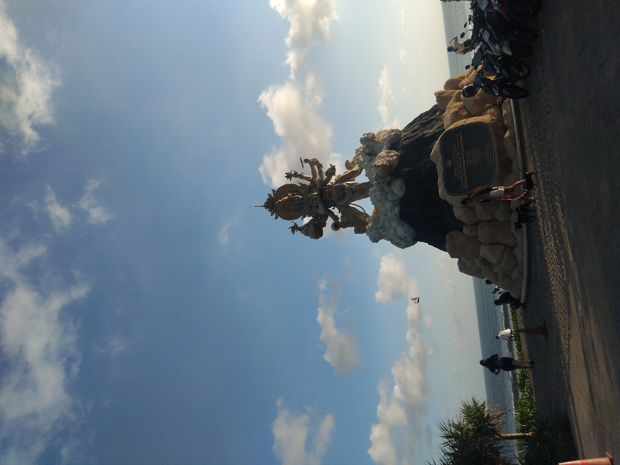 The image shows a statue of a multi-armed deity on a rocky base, set against a backdrop of a bright blue sky with scattered white clouds. The statue is positioned near a coastal area, with the sea visible in the background. The location seems to be a public area, with people walking by and several motorcycles parked nearby. There is also a stone plaque on the base of the statue.