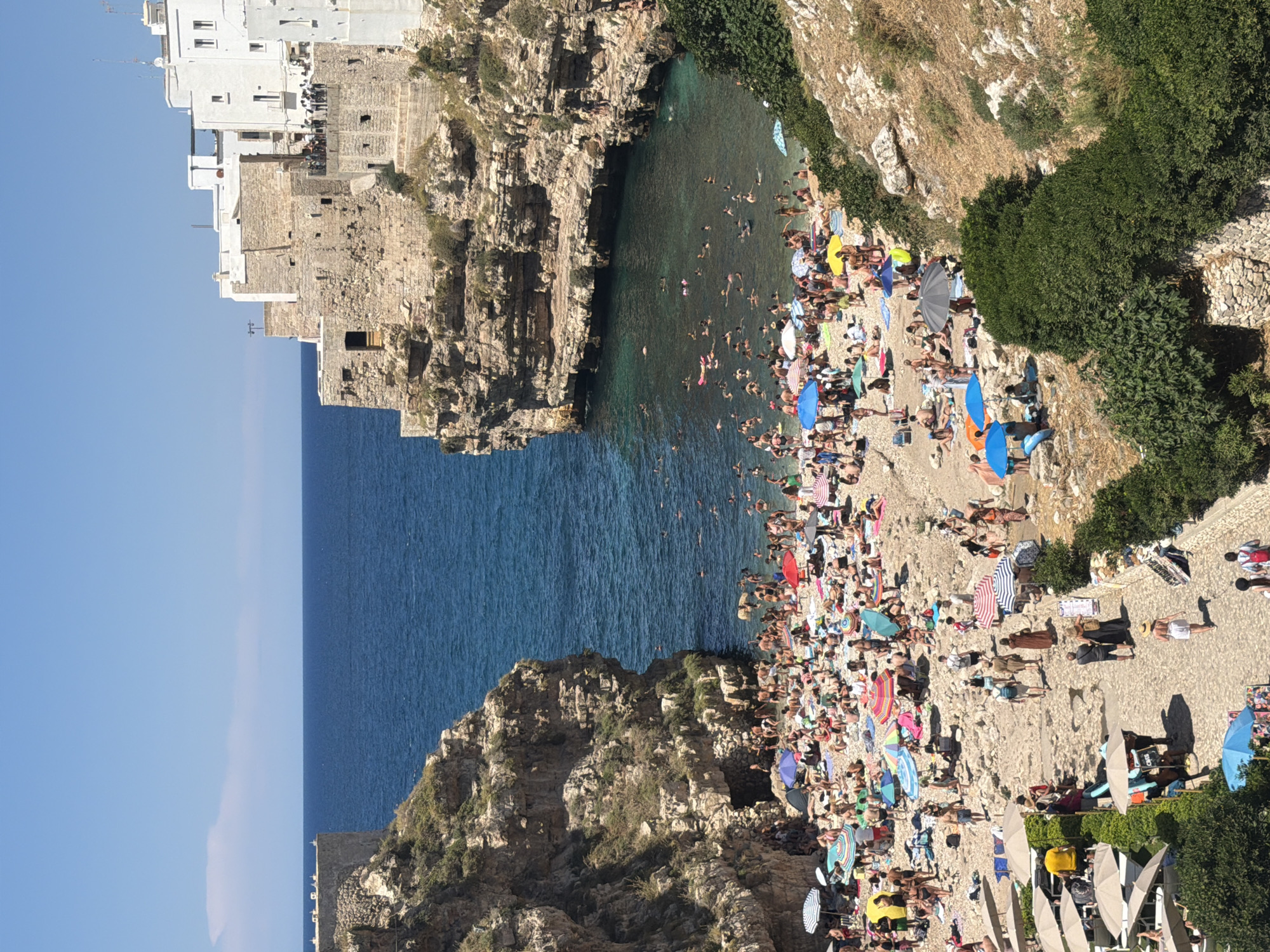 The image shows a crowded beach in Polignano a Mare, Italy, nestled between rocky cliffs. The water is a clear turquoise, and many people are swimming or floating in it. The beach itself is covered in pebbles, and numerous umbrellas are set up, providing shade for sunbathers. People are scattered across the beach, some lounging on towels, others walking or interacting with each other. Buildings, characteristic of Polignano a Mare's architecture with white facades and stone accents, are perched on the cliff tops overlooking the bay. The sky is clear and blue, indicating a sunny day. Lush green vegetation grows on the surrounding cliffs.