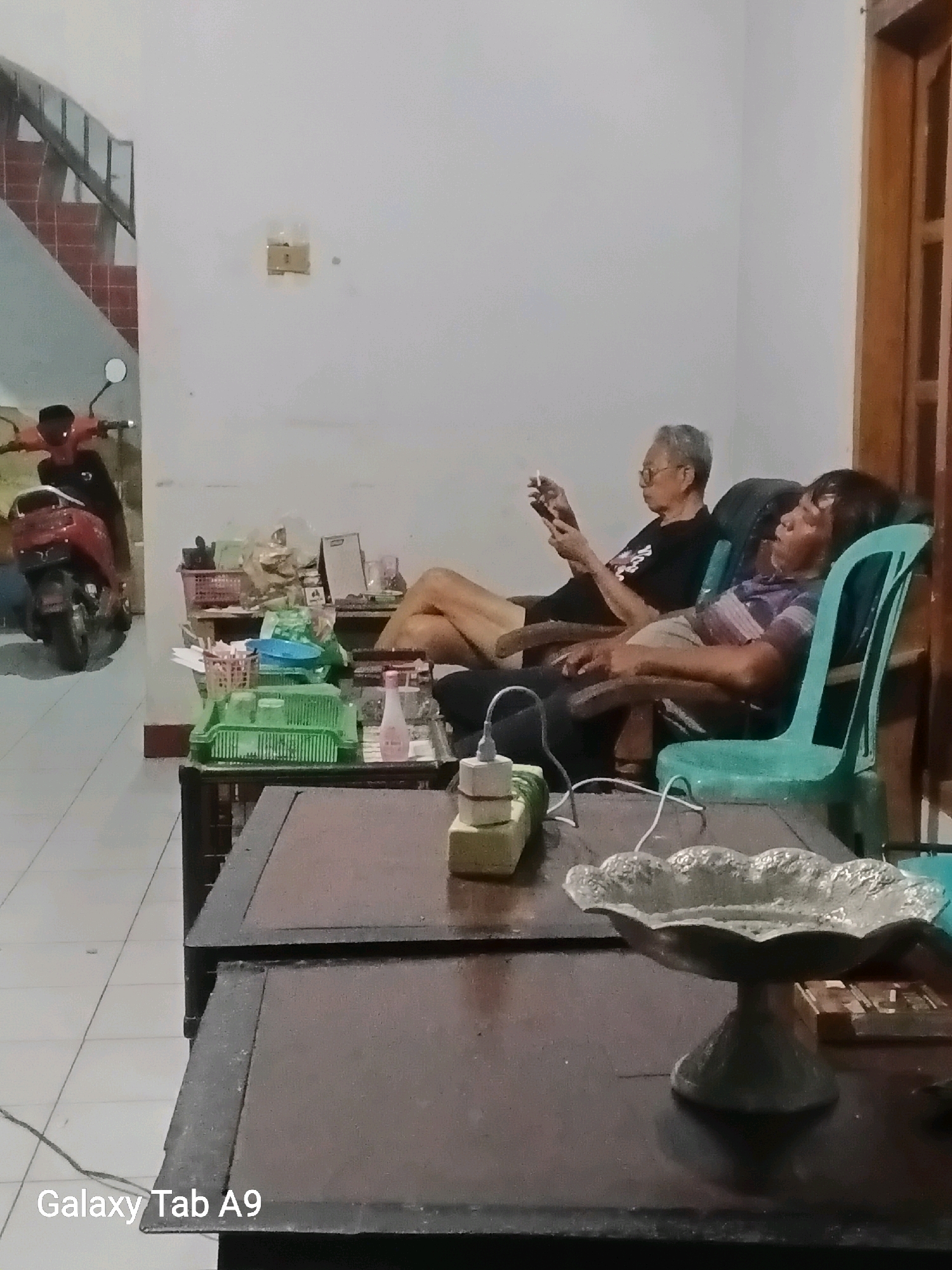 Two individuals are seated in chairs in a living room. The person on the left, a man with gray hair and glasses, is holding a smartphone and appears to be smoking a cigarette. He is wearing a black t-shirt. The person on the right, a man with dark hair, is leaning back in a teal plastic chair and appears to be asleep or resting, with his mouth slightly open. He is wearing a striped t-shirt. In the foreground, a dark wooden table holds various items, including a decorative metal bowl, a power strip with cords, and a green plastic basket containing utensils. Behind the table, to the left, is a red scooter. The room has white walls, tiled flooring, and a staircase with red steps and a metal railing in the background. The overall scene suggests a casual, indoor setting.