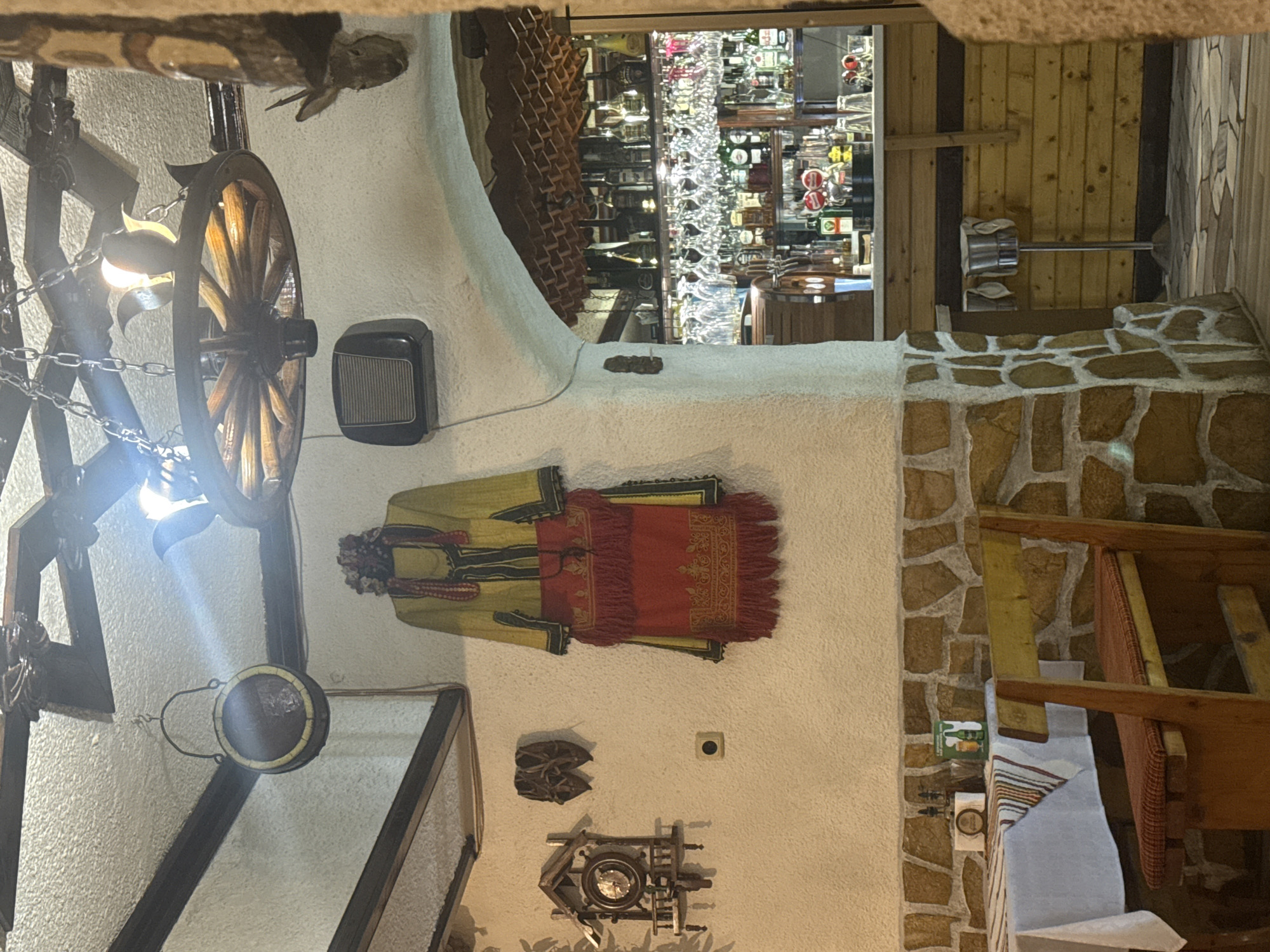 The image shows the interior of a restaurant, likely in Varna, Bulgaria, given the traditional Bulgarian clothing and decorative elements. The interior has textured white walls with exposed stone accents. A wagon wheel chandelier hangs on the left. A traditional Bulgarian costume is displayed on the wall. A small display case, shoes, and an antique machine are also mounted on the wall. A doorway in the background reveals a bar with a variety of bottles and glasses. Wooden beams are also visible around the opening. On the right, there is furniture with white tablecloths and stone walls.