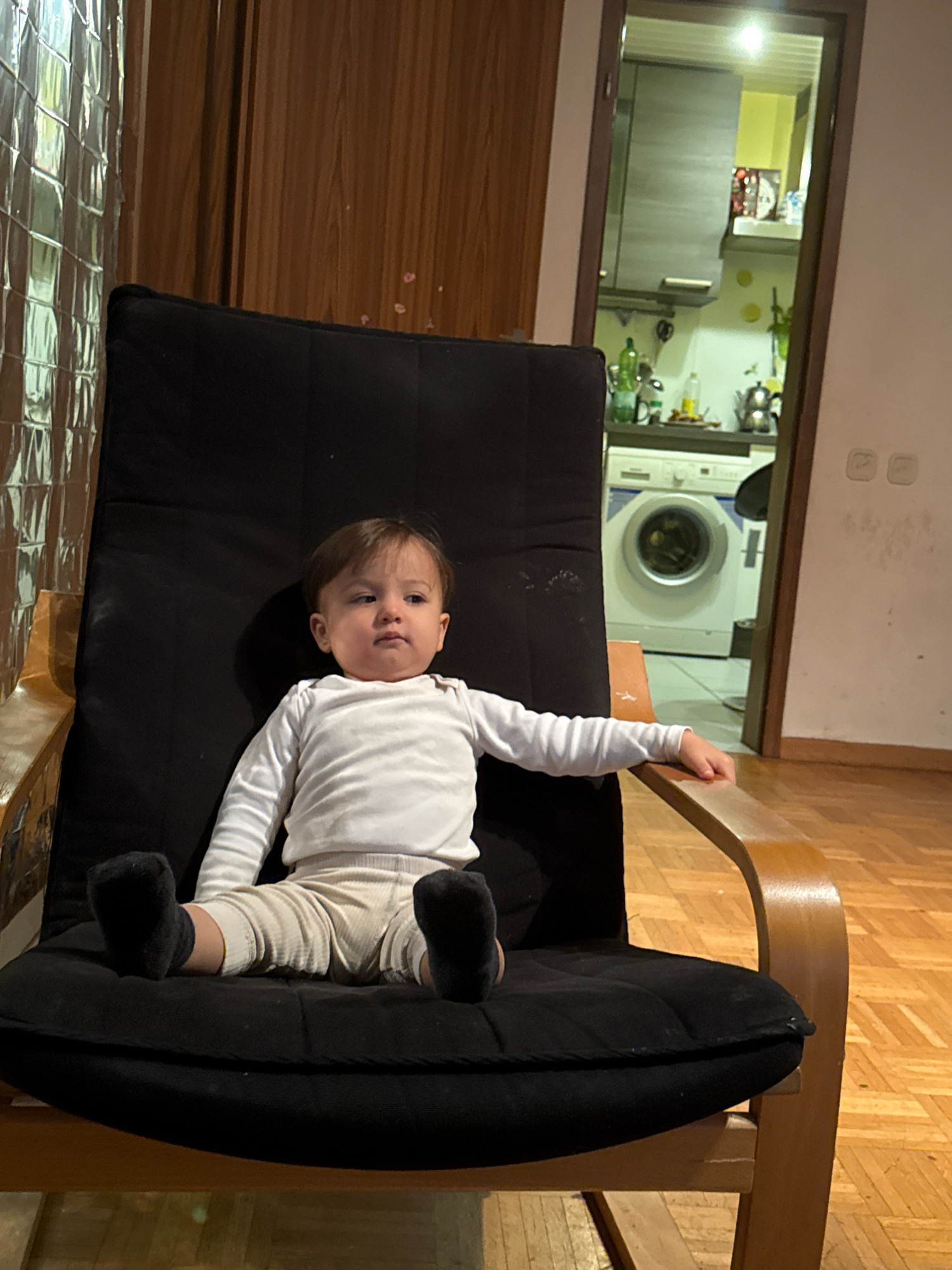 A young child is sitting in a black upholstered chair with wooden arms and legs. The child is wearing a white long-sleeved shirt and beige pants, with black socks on their feet. The child is looking slightly to the right with a neutral expression. Behind the child, there is a doorway leading to a kitchen area where a washing machine is visible. The flooring appears to be wood. The overall scene depicts a domestic indoor environment. The location is Überherrn, Germany.