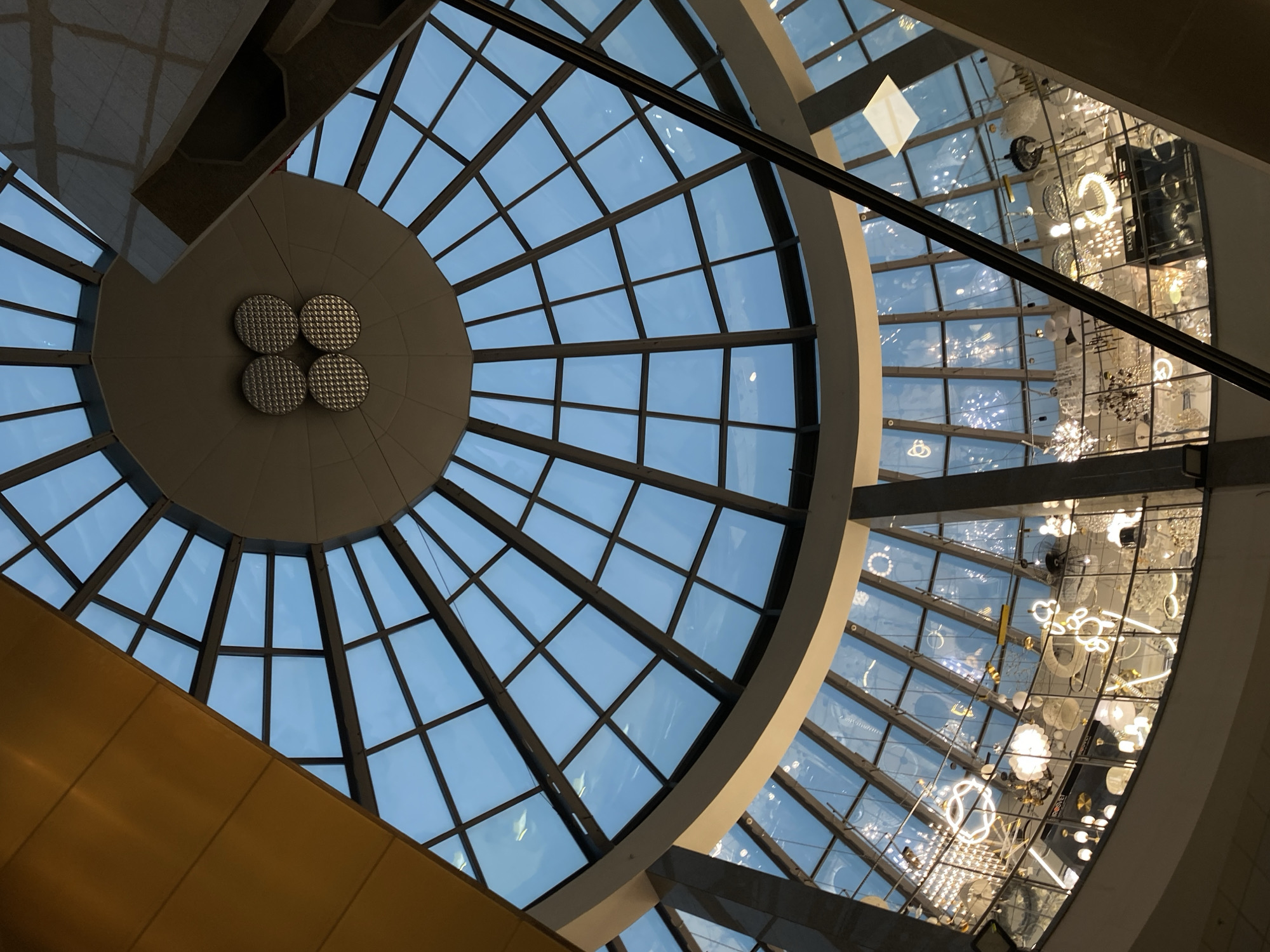 This image depicts an upward view inside a building, possibly a shopping center or mall. The architectural design features a large, circular glass ceiling, reminiscent of a dome, with a radial pattern of panes divided by a dark support structure. The sky is visible through the glass, indicating daylight. To the right, another section of glass roof, possibly part of a different wing or section of the building, showcases an array of modern lighting fixtures hanging from the ceiling. There's a bar running across the image. There is also a top section of ceiling on the right-hand side of the picture. The image's angle suggests it was taken from a lower level, looking upwards towards the roof structures. The city is Kazan, Russia.