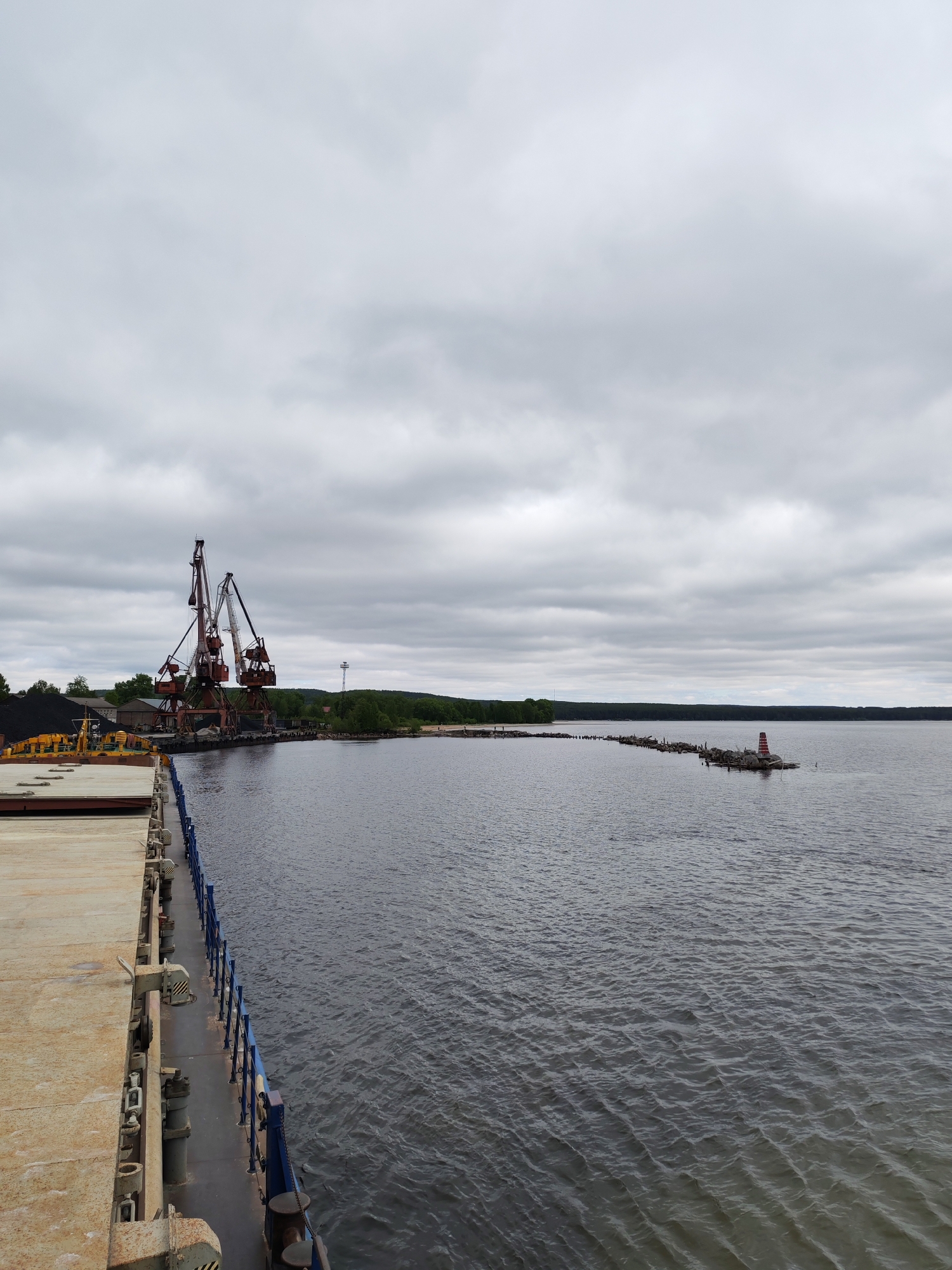 The image captures an industrial port scene on a cloudy day in Medvezhyegorsk, Russia. In the immediate foreground on the left, the weathered and rusty deck of a ship or barge is visible, featuring a blue railing that extends along its side, suggesting the vessel is docked or moving alongside a waterfront.
In the midground to the left, two prominent industrial port cranes, characterized by their rusty reddish-brown and grey steel structures, stand at the dock. Adjacent to the cranes, a large pile of dark material, likely coal, is visible, indicating cargo handling operations. Behind the dock area, a few low buildings are nestled among green trees.
The center and right side of the image are dominated by a wide body of dark water, showing subtle ripples across its surface. Extending from the distant, forested shoreline into the water is a long, low structure composed of logs or wooden pilings. At the far end of this structure, a distinct red and white striped navigational marker or small lighthouse stands, aiding in waterway navigation. The background features a continuous stretch of low, tree-covered land along the horizon, all set beneath a uniform, overcast grey sky with diffuse clouds.
The scene conveys a quiet, working port environment. No individuals are visible, and no specific activity is explicitly taking place. The time of day appears to be daytime, under heavily overcast conditions. No discernible text is present in the image.
