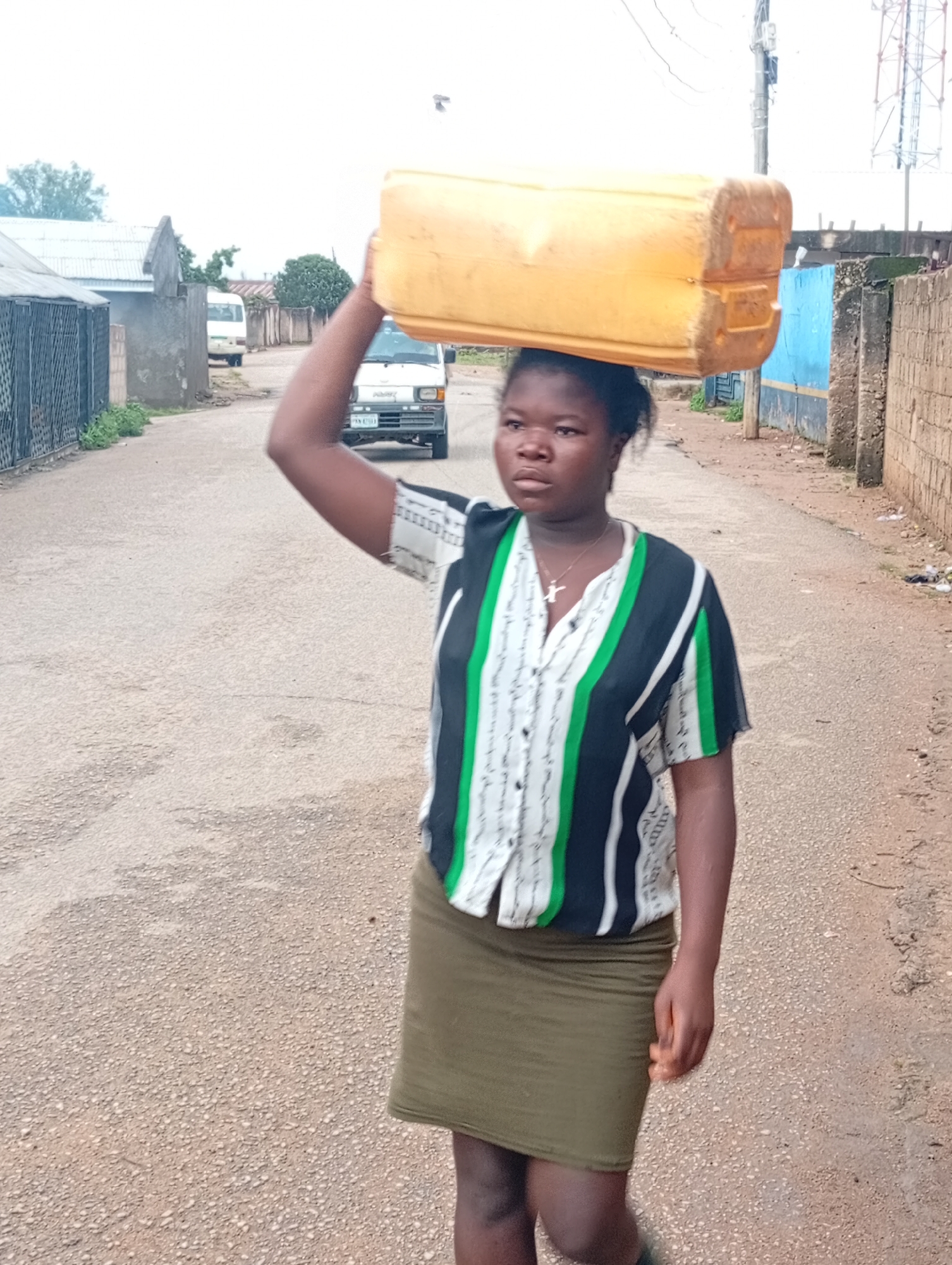 A young person, identified as a female, walks along an asphalt road, carrying a large yellow plastic container of water balanced on her head. She is wearing a vertically striped shirt with black, green, and white colors, and a green skirt. The environment appears to be a residential area in Jos, Nigeria, with houses, fences, and a telephone pole visible in the background. A white van is parked on the side of the road. The sky is overcast.