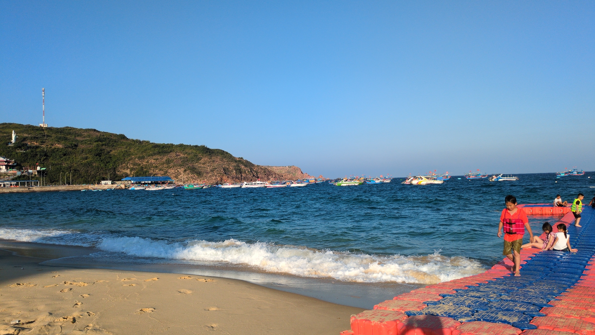 The image presents a daytime coastal scene in East Quy Nhon Ward, Vietnam, under a clear blue sky. The foreground features a light brown sandy beach with visible footprints, meeting the ocean where small white waves break. Extending from the beach into the water on the right is a modular floating platform constructed from interlocking red and blue plastic sections.
On this platform, a young male, appearing to be a child, walks barefoot on the blue section, wearing a red life vest and dark shorts, facing left. Behind him, two younger children sit on the blue platform, one in a white top, facing right, while another child wearing a yellow-green life vest stands further back on the platform. Other individuals are partially visible on the platform and in the water further out.
The middle ground is dominated by the calm, blue-green ocean surface, populated by numerous boats of various sizes and colors, including white speedboats, green and blue passenger boats, and several fishing vessels, either anchored or navigating the bay.
To the left, a prominent green, tree-covered hill rises, featuring a white statue on its slope, a red-roofed building, and a pier extending from its base into the water. At the summit of the hill, a tall, white and red communication tower is visible. The scene is illuminated by bright sunlight.