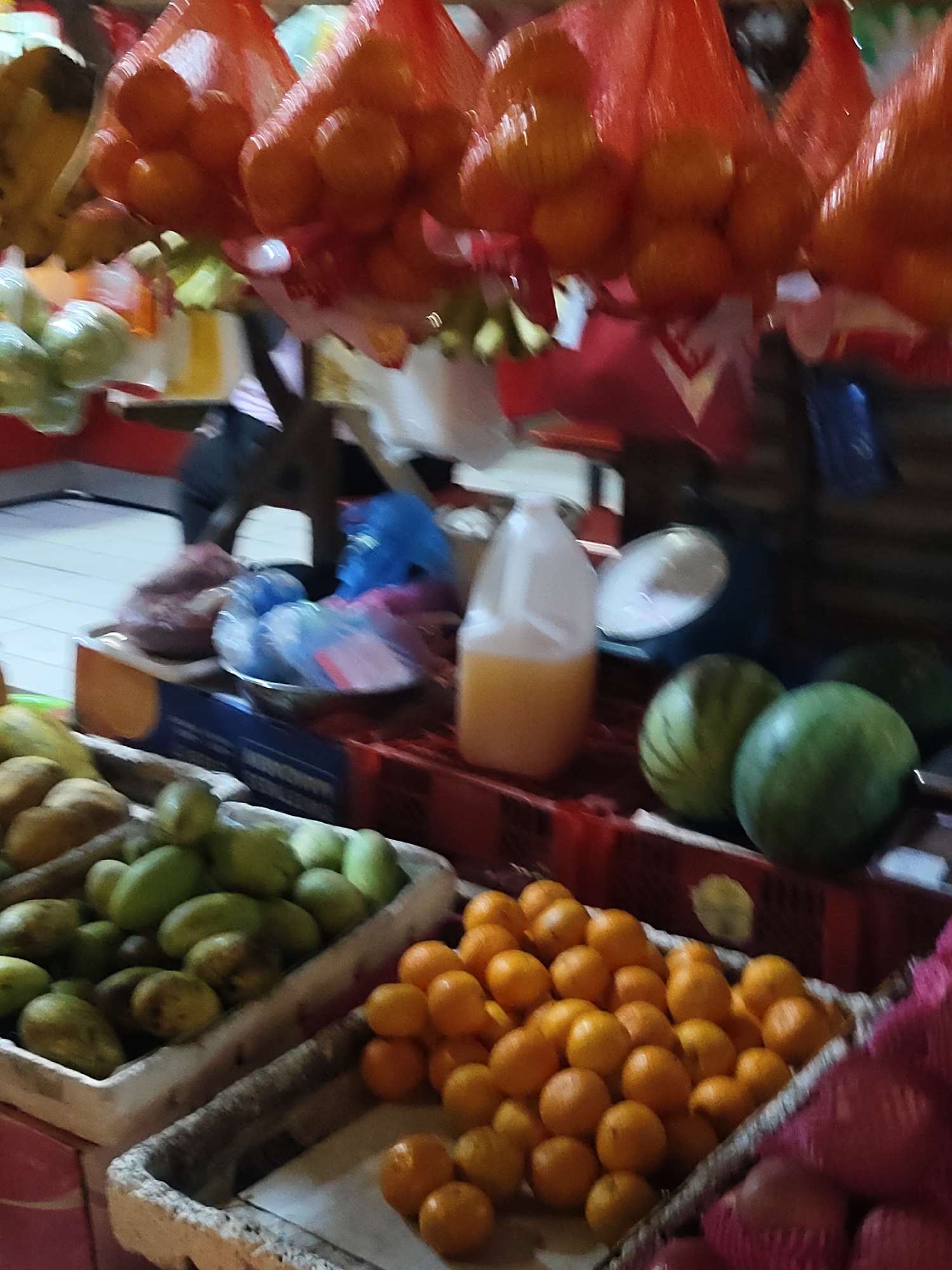 The image depicts a vibrant market scene, likely in Davao City, Philippines, showcasing a variety of fresh produce. In the foreground, several crates are filled with different fruits. One crate contains numerous small, orange citrus fruits, possibly mandarins or clementines, neatly arranged. Adjacent to it, another crate holds an assortment of green, elongated fruits, which appear to be unripe mangoes or similar tropical produce. Further back, two watermelons with distinctive stripes are visible, alongside other green fruits.
Suspended from above are several red mesh bags filled with vibrant orange fruits, likely oranges or tangerines. These are a prominent feature in the upper part of the image. In the middle ground, a large translucent jug filled with a yellowish liquid, possibly juice, sits on a red tiered display. Behind the jug, a metal scale is partially visible, suggesting a point of sale or weighing station.
The background is somewhat blurred but suggests the bustling environment of an outdoor or semi-enclosed market. Wrapped items, possibly meat or other goods, are visible in plastic bags, placed in containers. The lighting suggests it could be daytime, as is typical for market operations. The overall impression is one of abundance and the lively atmosphere of a local fruit market. There is no visible text in the image.