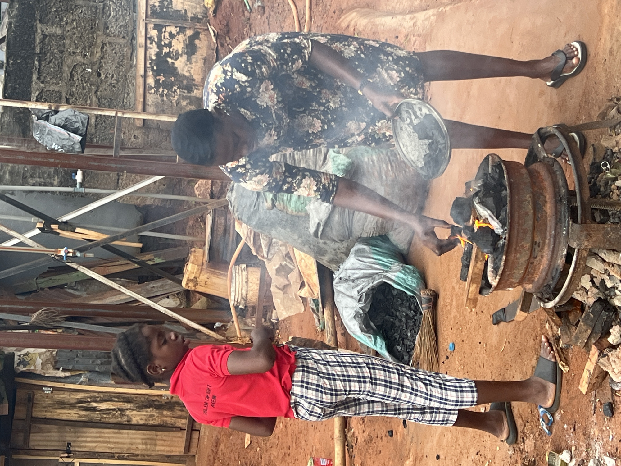 In Nkpor, Nigeria, two young Black individuals are engaged in an activity around a makeshift outdoor fire. The scene is set on reddish-brown earth, with a backdrop of rudimentary structures constructed from corrugated metal, wood, and what appear to be concrete blocks. A pile of charcoal is visible in a large, green-and-gray sack to the left of the frame, with a broom leaning against it.
In the foreground, a younger Black child, wearing a red t-shirt and checkered pants, stands facing the fire, looking down. The t-shirt has lettering on the back, though it is partially obscured. A slightly older Black individual, wearing a dark floral-patterned dress, is positioned to the right, actively tending to the fire. They are holding a shallow metal dish, from which they are adding charcoal to the flames. The fire is contained within a circular, metal apparatus that appears to be a repurposed vehicle rim, elevated on a stand made of scrap metal. The ground around the fire is scattered with wood and charcoal. The overall environment suggests a practical, outdoor setting, likely a residential or working area.
