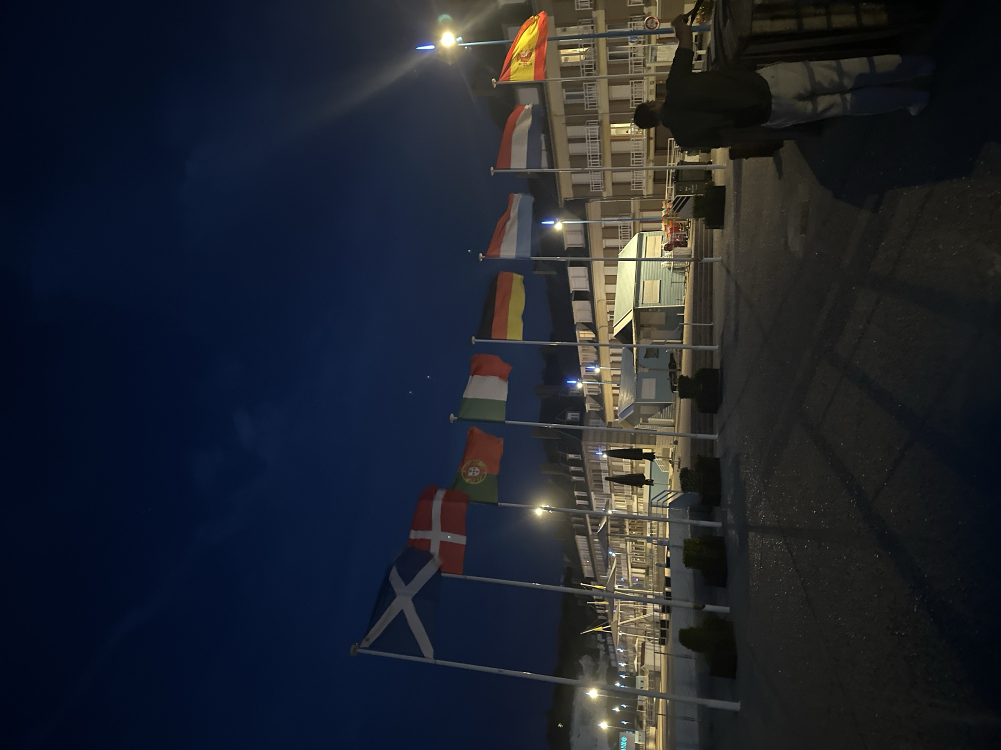 The image captures a view of the waterfront in Saint-Valery-en-Caux, France, at night. A line of flags, including those of Scotland, Denmark, Portugal, Italy, France, Germany, Luxembourg, the Netherlands, and Spain, are flying from poles along the promenade. In the foreground, a person is standing near a dark wooden structure, facing away from the camera and towards the water. Behind the flags, a row of buildings with illuminated windows lines the waterfront. The sky is dark blue with some faint clouds. Streetlights and building lights illuminate the scene.
