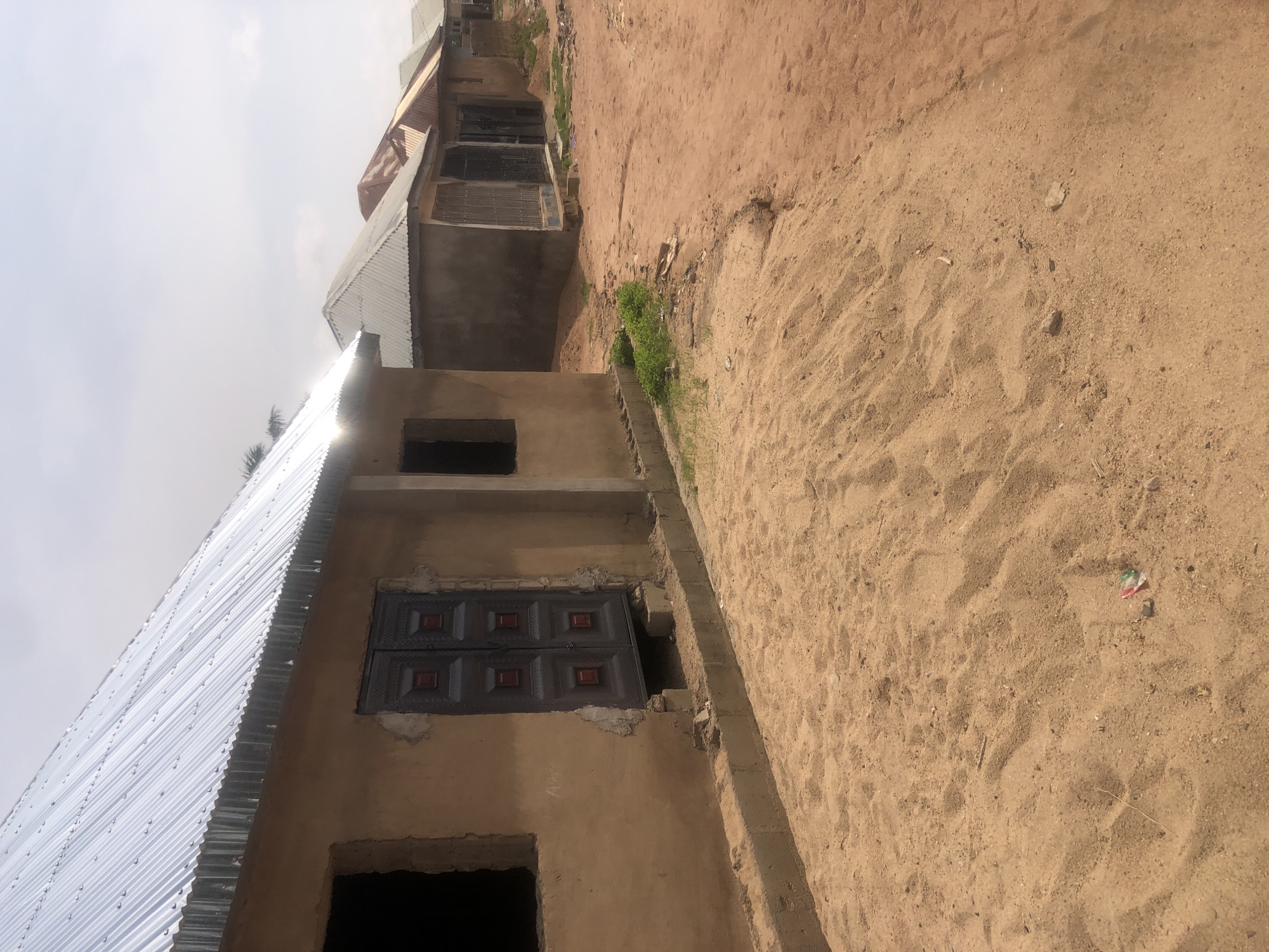 In Jalingo, Nigeria, a row of unpainted buildings with corrugated metal roofs line a sandy path. The buildings appear to be under construction, with some having open window frames and exposed concrete foundations. One building in the foreground has a dark, double-doored entrance with red accents. The ground is covered in light brown sand, with sparse green vegetation growing along the edges of the buildings. The sky is overcast, with a few faint clouds visible. A small piece of colorful trash is visible in the sand in the foreground.