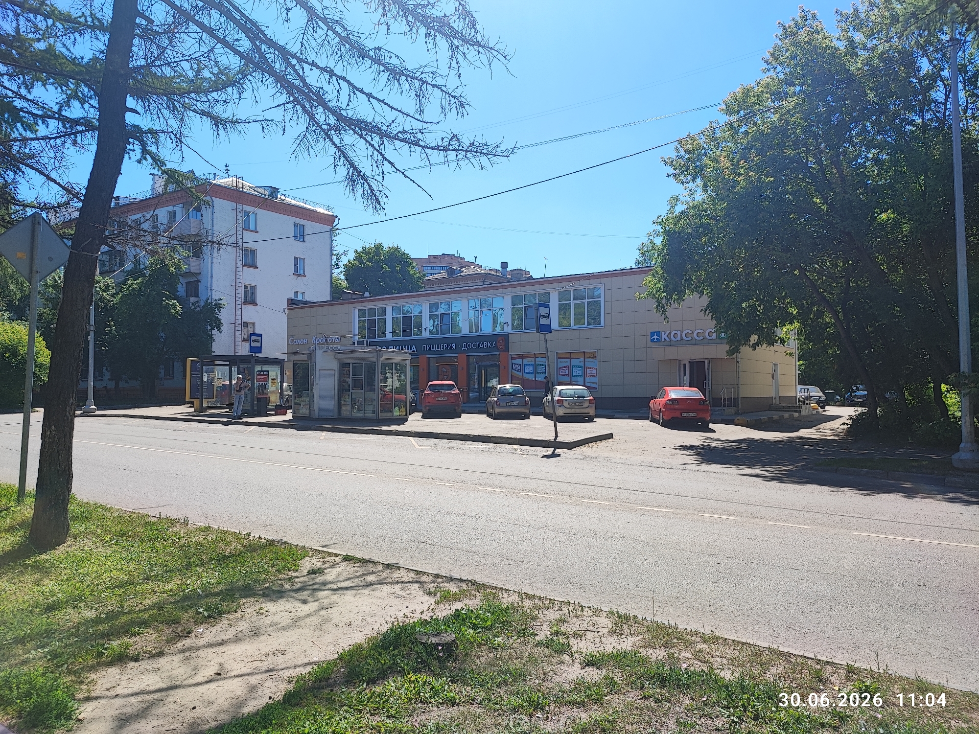 This is a daytime, eye-level, full outdoor shot of a street scene in Lytkarino, Russia. In the foreground, a paved road with a white dashed line runs horizontally across the image. To the left of the road is a grassy embankment with a dirt path and a tree trunk on the far left. In the mid-ground, there is a bus stop shelter with advertisements and a few people standing near it. A white and yellow single-story building with large windows is visible behind the bus stop. This building has signage that appears to be in Russian, with one sign reading "Касса" (Kassa), indicating a ticket office or cash desk. Several cars are parked in front of the building, including a red sedan, a silver hatchback, and a white hatchback. In the background, a multi-story apartment building with white and orange detailing stands tall. Lush green trees are prevalent throughout the scene, particularly on the right side of the image, casting shadows on the road and sidewalk. The sky is clear and blue. A timestamp in the bottom right corner reads "30.06.2026 11:04".