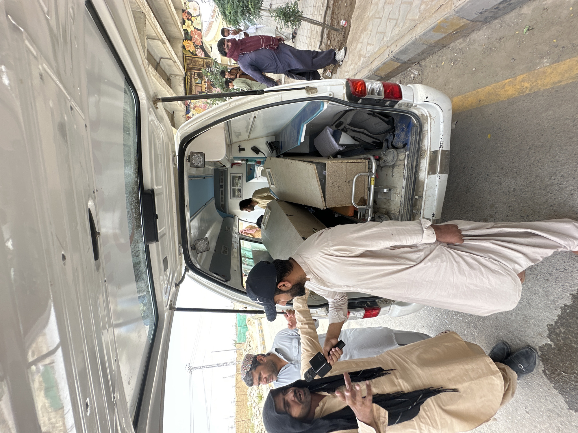 A low-angle, wide shot captures the interior and exterior of a white ambulance parked on a street in Quetta City Tehsil, Pakistan. The rear doors of the ambulance are open, revealing a stretcher with a wooden box on it, a metal shelf unit, and bags. Two men are standing near the open doors. One man, wearing a tan shalwar kameez and a black headscarf, is holding a microphone and appears to be speaking to the other man, who is wearing a tan shalwar kameez and a black baseball cap. Another man in a light blue shalwar kameez stands behind them, looking to the left. In the background, several other men are visible, one wearing a dark blue shalwar kameez and a burgundy scarf covering his face. The street appears to be paved with asphalt, with a yellow and gray curb on the right side. The environment is somewhat dusty and unkempt, with construction materials visible in the distance.