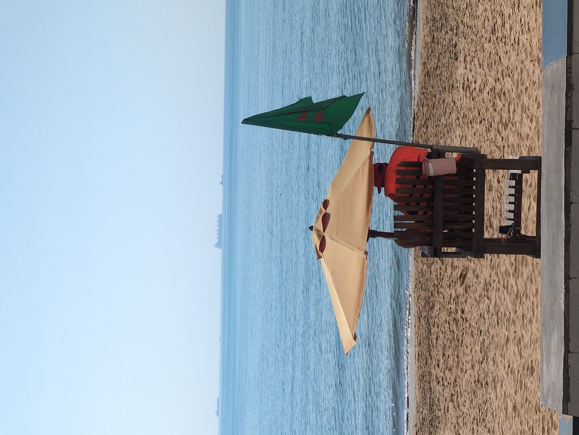 The image depicts a beach scene in South Quy Nhon Ward, Vietnam, under a clear, light blue sky. A lone individual, appearing to be a lifeguard, is seated on a elevated wooden lifeguard chair, facing the calm blue ocean. The person is wearing a prominent orange life vest or uniform, their back visible to the viewer. A beige circular umbrella provides shade over the chair. To the right of the umbrella, a green flag with red lettering (likely Vietnamese characters) is affixed to a pole, extending upwards from the lifeguard station.
The sandy beach occupies the mid-ground, showing ripple patterns from wind or water, meeting the gentle waves of the sea. Several dark-hulled cargo ships are visible on the distant horizon, indicating a shipping lane or port nearby. The immediate foreground shows a low, grey concrete barrier or ledge. No other people or significant interactions are visible.