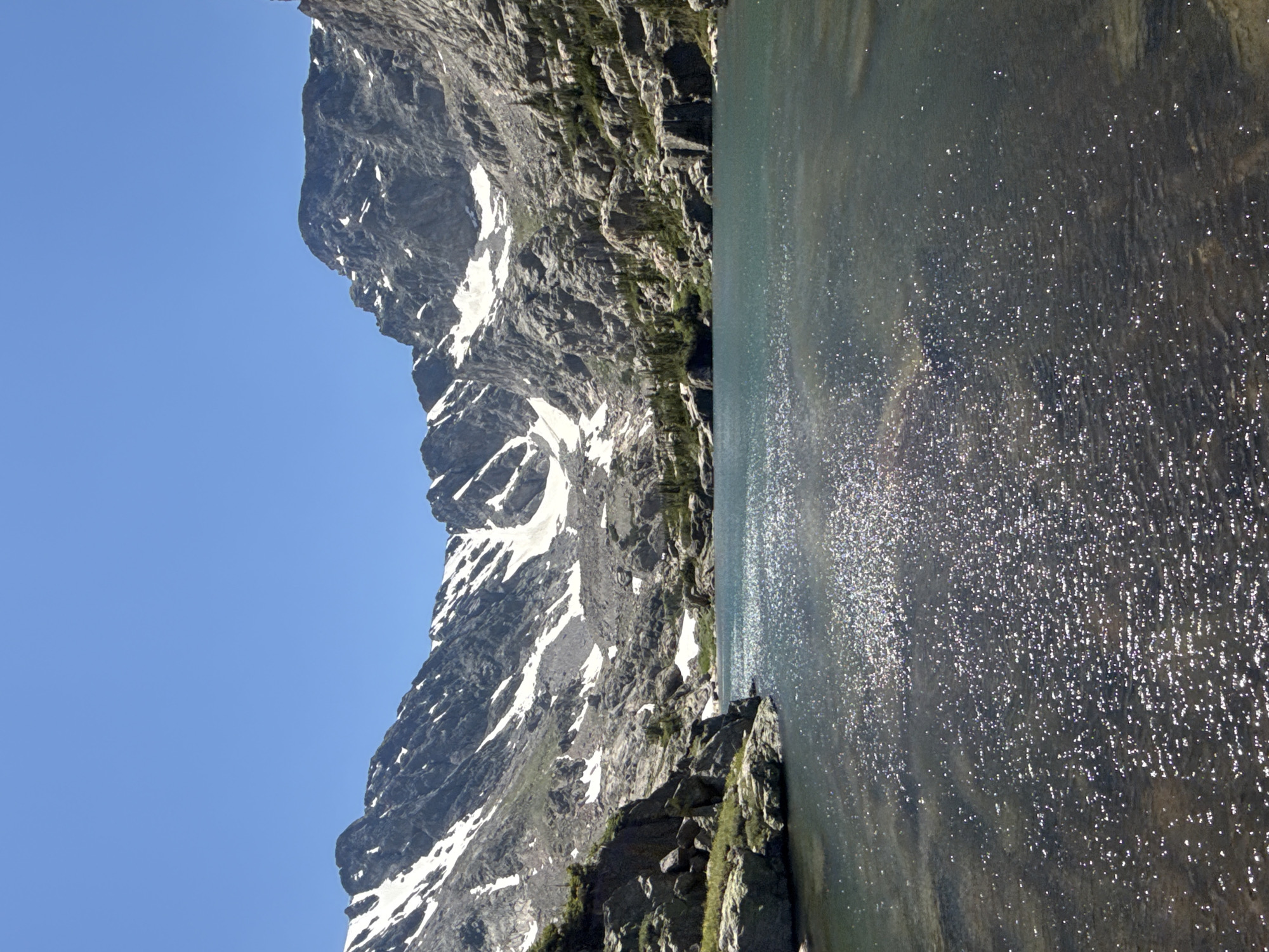 This is a vertical, eye-level, full shot of a mountain lake with snow-capped peaks in the background. The sky is clear and blue. The lake is a deep turquoise and sparkles with sunlight. The surrounding mountains are rugged and rocky, with patches of snow still visible. There are scattered evergreen trees growing on the lower slopes of the mountains. The foreground shows the edge of the lake with large rocks and some vegetation. The image appears to be taken on a sunny day in a natural, mountainous environment. The location is likely in the Rocky Mountains near Allenspark, Colorado, given the characteristic landscape. There are no people or man-made structures visible in the image.