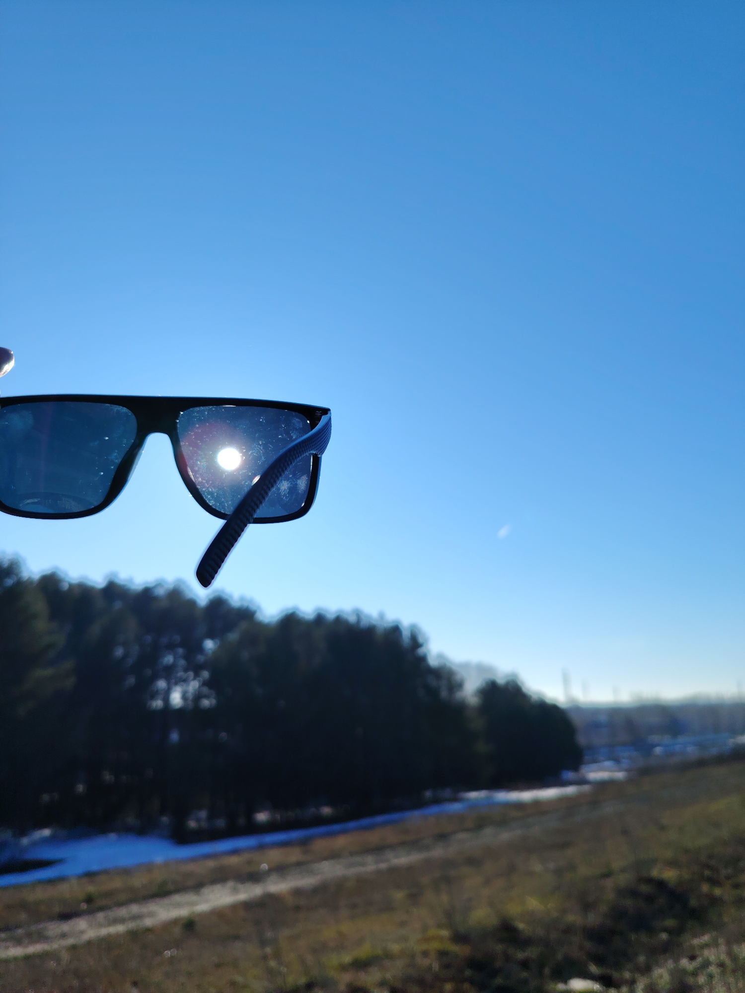 A photograph captures a pair of black-framed sunglasses being held up against a clear blue sky. The sun's reflection is visible on the lens of the sunglasses. Below the sunglasses, a blurred landscape unfolds, featuring a line of evergreen trees and a patch of remaining snow. In the far background, indistinct industrial structures can be discerned, suggesting the location might be near the city of Chaikovsky, Russia.