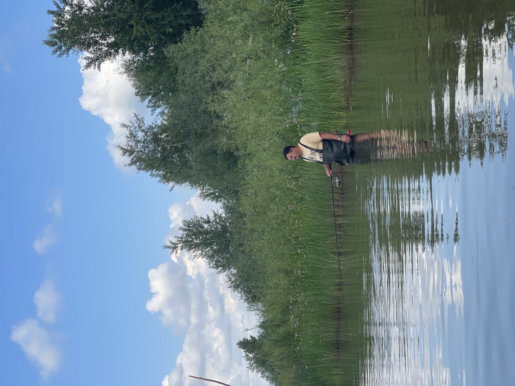 A man is standing in shallow water, wearing waders and holding a fishing rod. He is facing to the left, with a slight smile on his face. The water is calm, with reflections of the sky and surrounding vegetation. The background is filled with lush green reeds and trees, with a blue sky and white clouds above. The scene appears to be a natural, rural setting, possibly in the vicinity of Nezhinka, Russia, a village known for its natural beauty and agricultural landscapes. The man is engaged in fishing, a common recreational activity in such environments. The overall atmosphere is peaceful and serene, suggesting a pleasant day spent outdoors.