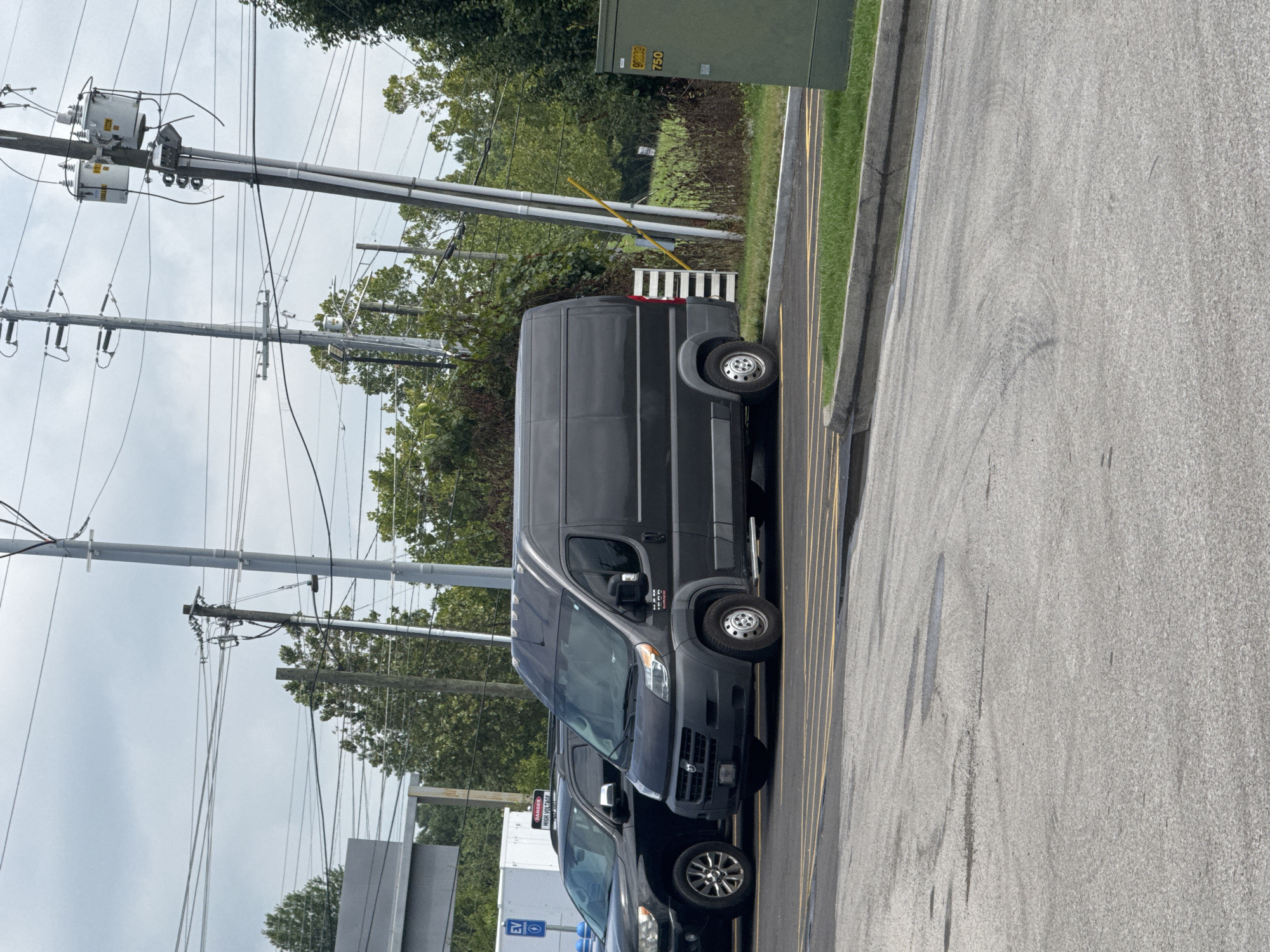 The image depicts an outdoor scene on an overcast day. In the foreground, a gray asphalt parking lot is visible, marked with yellow lines. To the right, a tall, green electrical transformer box stands. Behind the transformer box, there is a patch of green grass and dense foliage. Dominating the mid-ground is a dark gray cargo van parked in a parking space. To the left of the van, a dark-colored SUV is also parked. In the background, several utility poles with numerous electrical wires stretch across the frame. Trees with green foliage line the horizon. A white building with a blue sign that says "EV" is partially visible to the left of the SUV. The scene appears to be a parking area, possibly at a commercial or public facility, in Knoxville, United States of America.