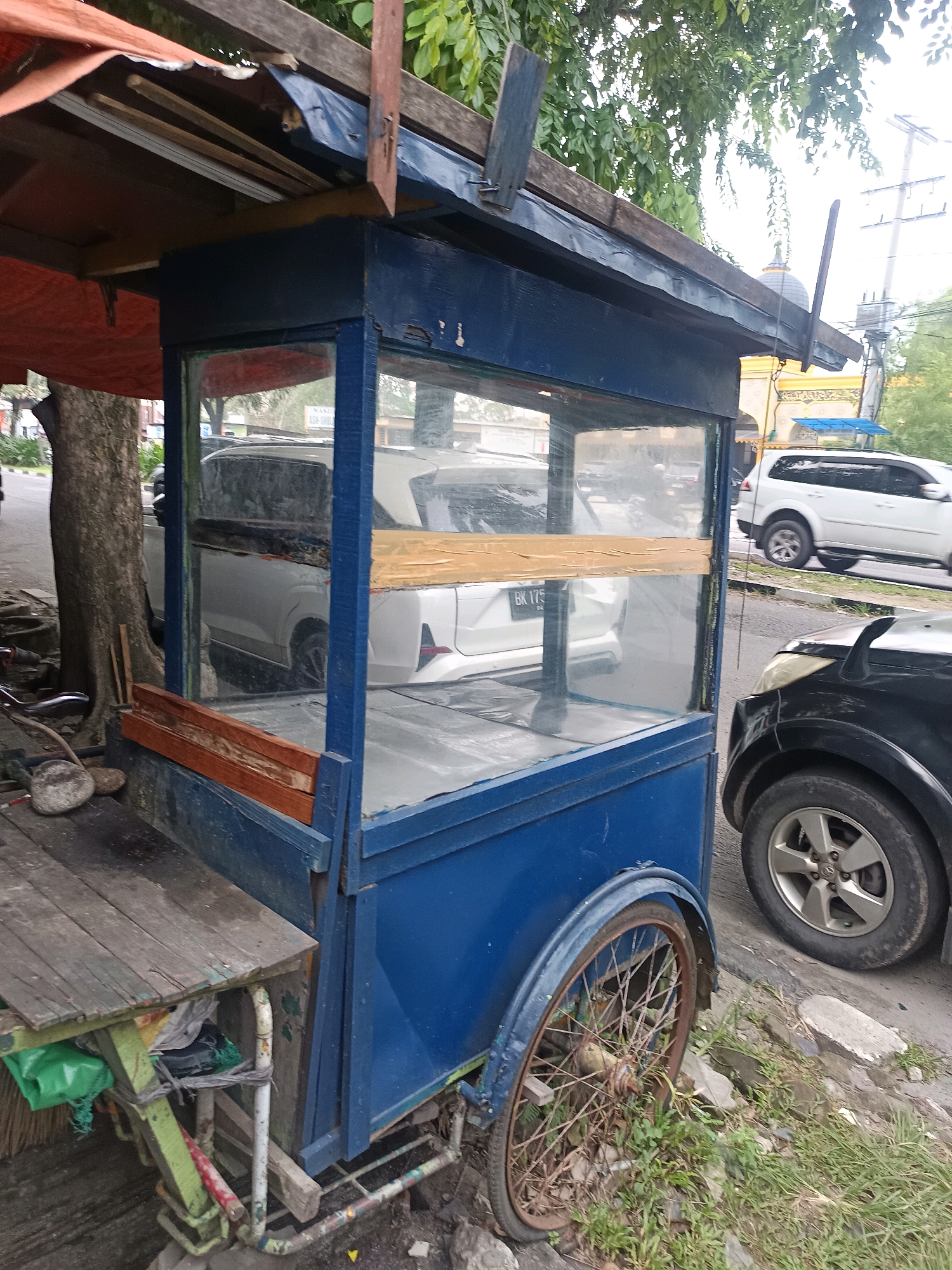 An outdoor scene in the city of Medan, Indonesia, depicts a weathered blue food cart with a wooden and metal frame. The cart has glass panels on its sides and front, offering a view into its interior which is mostly empty. A wooden table, showing signs of wear, is attached to the side of the cart. The cart has a single, spoked rear wheel. Behind the cart, several white SUVs are visible, suggesting a roadside location. A black car is parked to the right of the cart. The environment includes a large tree, grass, and some discarded items. The sky appears overcast.