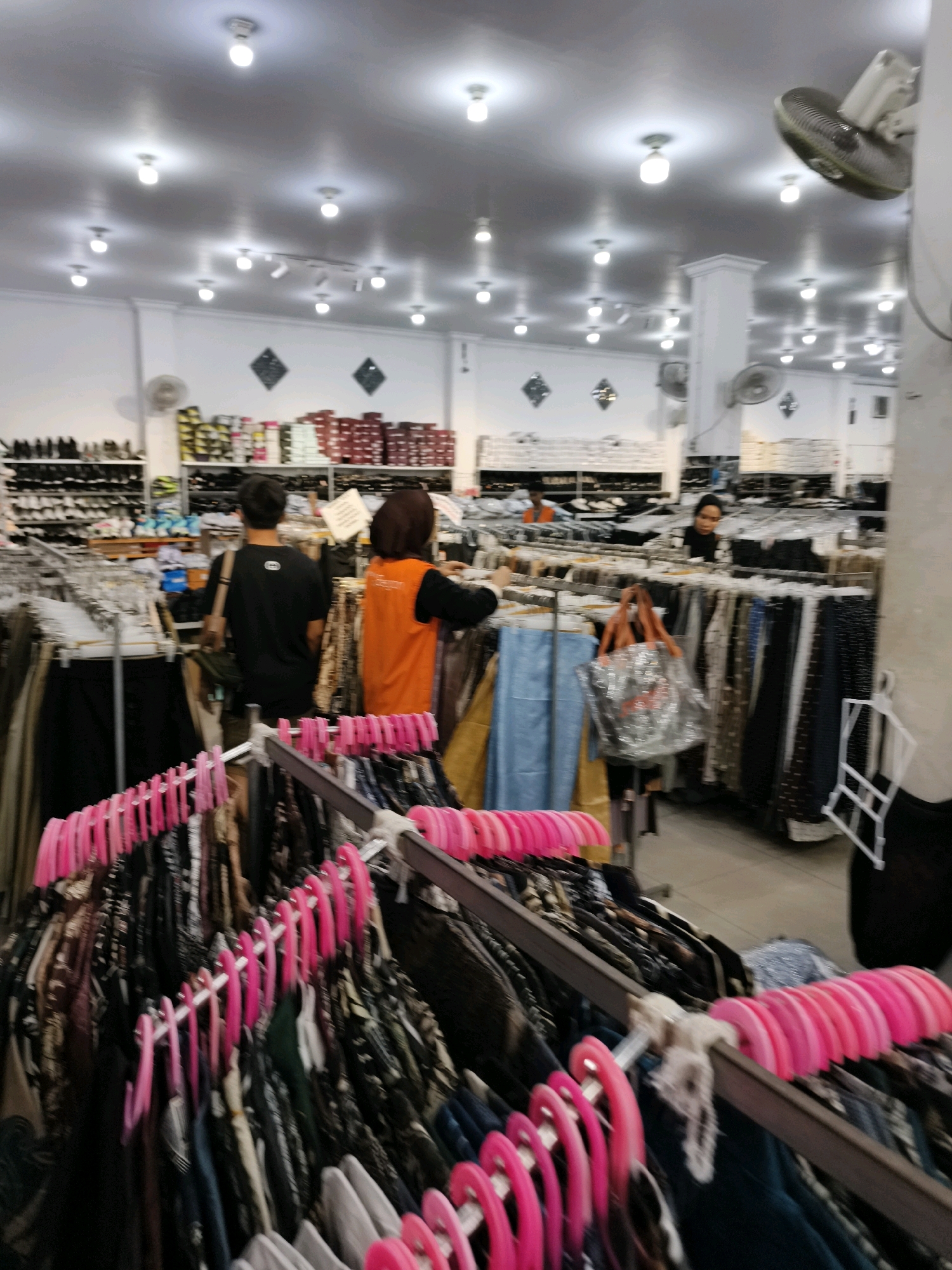 The image depicts the interior of a clothing store in Ketapang, Indonesia. In the foreground, rows of clothing on hangers with bright pink clips are visible, suggesting a retail environment. The store appears to be well-lit with numerous ceiling lights. In the mid-ground, several individuals are present. One person, wearing a black shirt, is facing away from the camera, looking towards the clothing racks. Another person, in an orange vest over a black garment, is also facing away and appears to be assisting a customer or arranging clothes. Further back, other shoppers and staff members can be observed browsing or working among the merchandise. Shelves stocked with various items, possibly footwear and more clothing, are visible along the walls. A ceiling fan is also present in the upper right portion of the image. The overall impression is that of a busy retail space selling apparel.