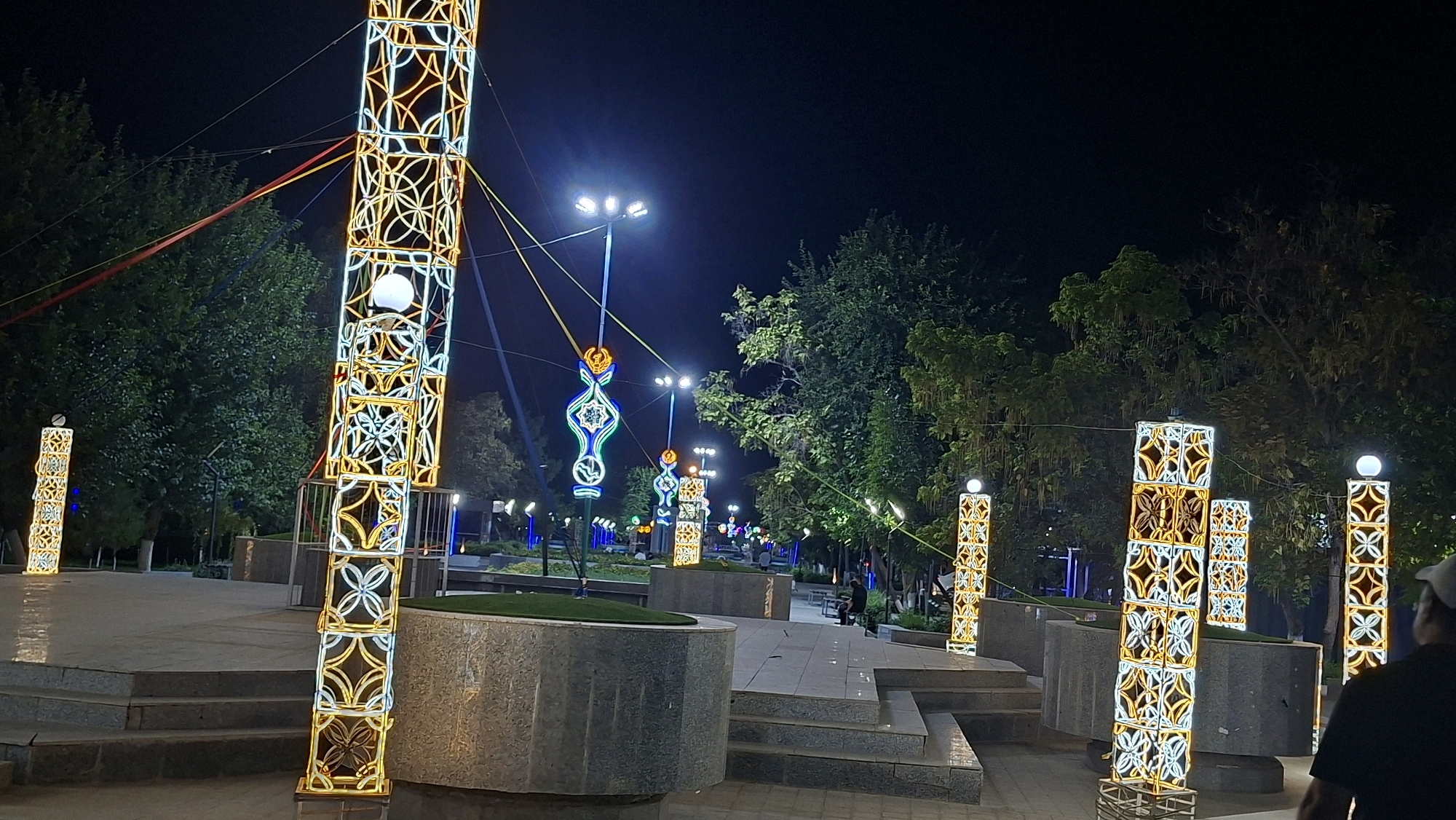 In the city of Navoiy, Uzbekistan, a park is illuminated at night by decorative, tall, rectangular pillars adorned with glowing geometric patterns of white and yellow lights. These pillars are spaced throughout the park, some standing individually and others grouped together. In the foreground, a person wearing a cap is visible from behind, looking towards the illuminated structures. In the background, trees with dark foliage form a backdrop against the night sky. Several streetlights with multiple bulbs are also present, casting a bright glow. Some of the decorative pillars also have spherical white lights at their apex. The ground is paved with light-colored tiles and concrete, with some stepped areas. In the mid-ground, more of these illuminated pillars and stylized light fixtures featuring green, blue, and yellow colors are visible, lining a path that leads into the park. The overall scene depicts a festive and well-lit urban park at night.