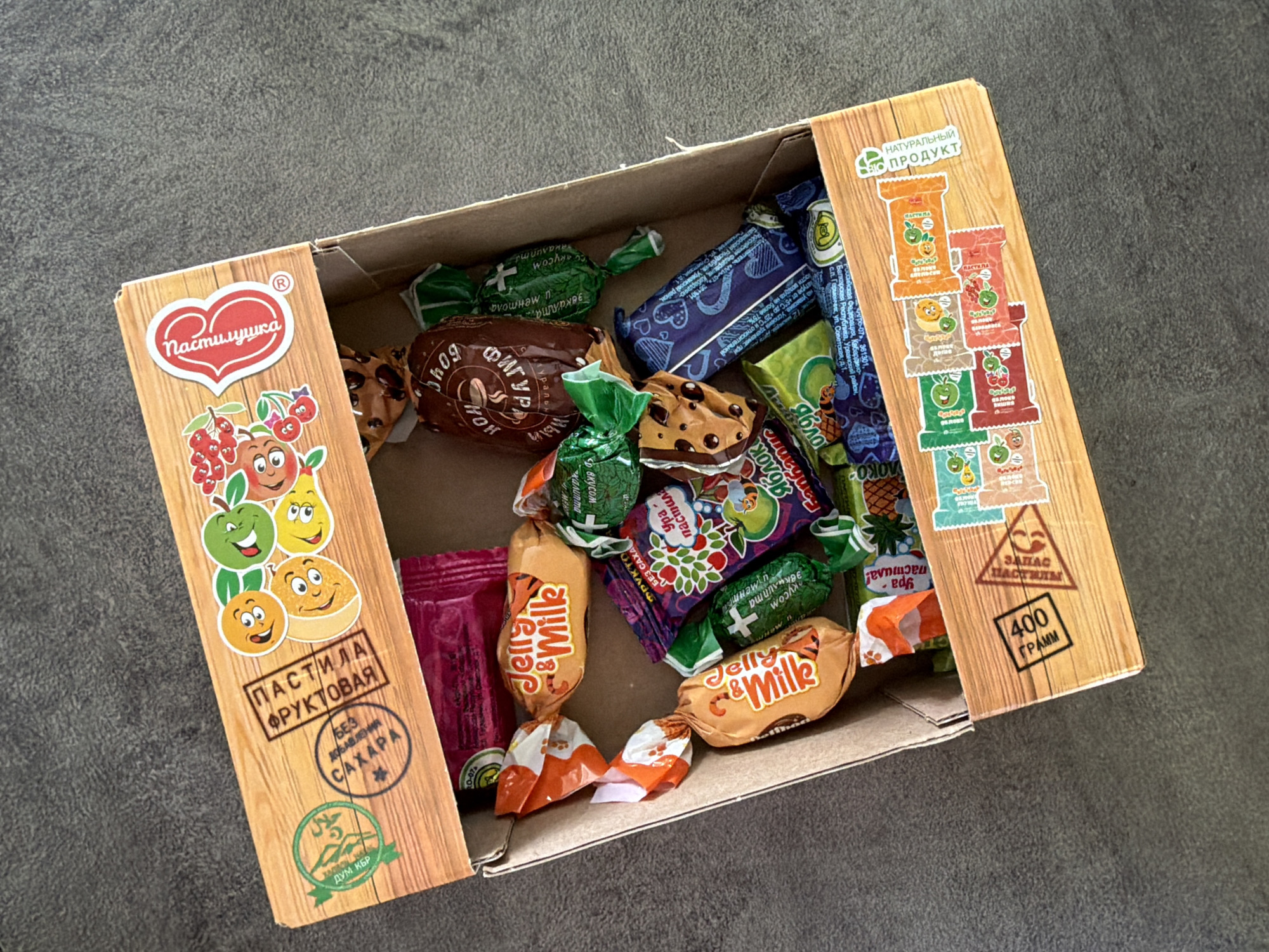 An overhead shot captures an open, light brown cardboard box filled with various individually wrapped candies, set against a dark, textured surface. The box is branded "Пастилушка" (Pastilushka), a Russian term for a type of fruit pastila.
The left inner flap of the box features a heart-shaped logo with "Пастилушка," below which are cheerful cartoon fruits—cherries, an apple, a pear, and an orange—suggesting the product's fruit base. Text on this flap reads "ПАСТИЛА ФРУКТОВАЯ" (Fruit Pastila) and "БЕЗ ДОБАВЛЕНИЯ САХАРА" (No Added Sugar), indicating a healthier option. A green circular logo with "ДУМ КБР" (Spiritual Administration of Muslims of Kabardino-Balkaria Republic) signifies Halal certification, confirming the product's Russian origin and market.
The right inner flap displays "НАТУРАЛЬНЫЙ ПРОДУКТ" (Natural Product) at the top. Below this, a column of small illustrations showcases different flavors of pastila in their wrappers, such as apple and cherry. Further down, a triangular stamp reads "ЗАПАС ПАСТИЛЫ" (Pastila Supply), and a box states "400 ГРАММ" (400 Grams), indicating the net weight.
Inside the box, a variety of colorful sweets are visible. Several "Jelly & Milk" candies in orange, tiger-striped wrappers are prominent, some partially unwrapped. Green-wrapped candies are scattered throughout, with visible text like "эвкалипта и ментола" (eucalyptus and menthol) and "Яблок" (Apple), suggesting fruit and medicinal flavors. One wrapper says "Ура пастила!" (Hurrah pastila!). A large brown candy wrapper features coffee bean graphics and the word "ФИГУР" (Figure). A blue wrapper with heart patterns, and a small pink/red packet, also add to the assortment. The presence of Cyrillic text across all branding and packaging confirms the product is intended for the Russian-speaking market, consistent with a location such as Chaikovsky, Russia.