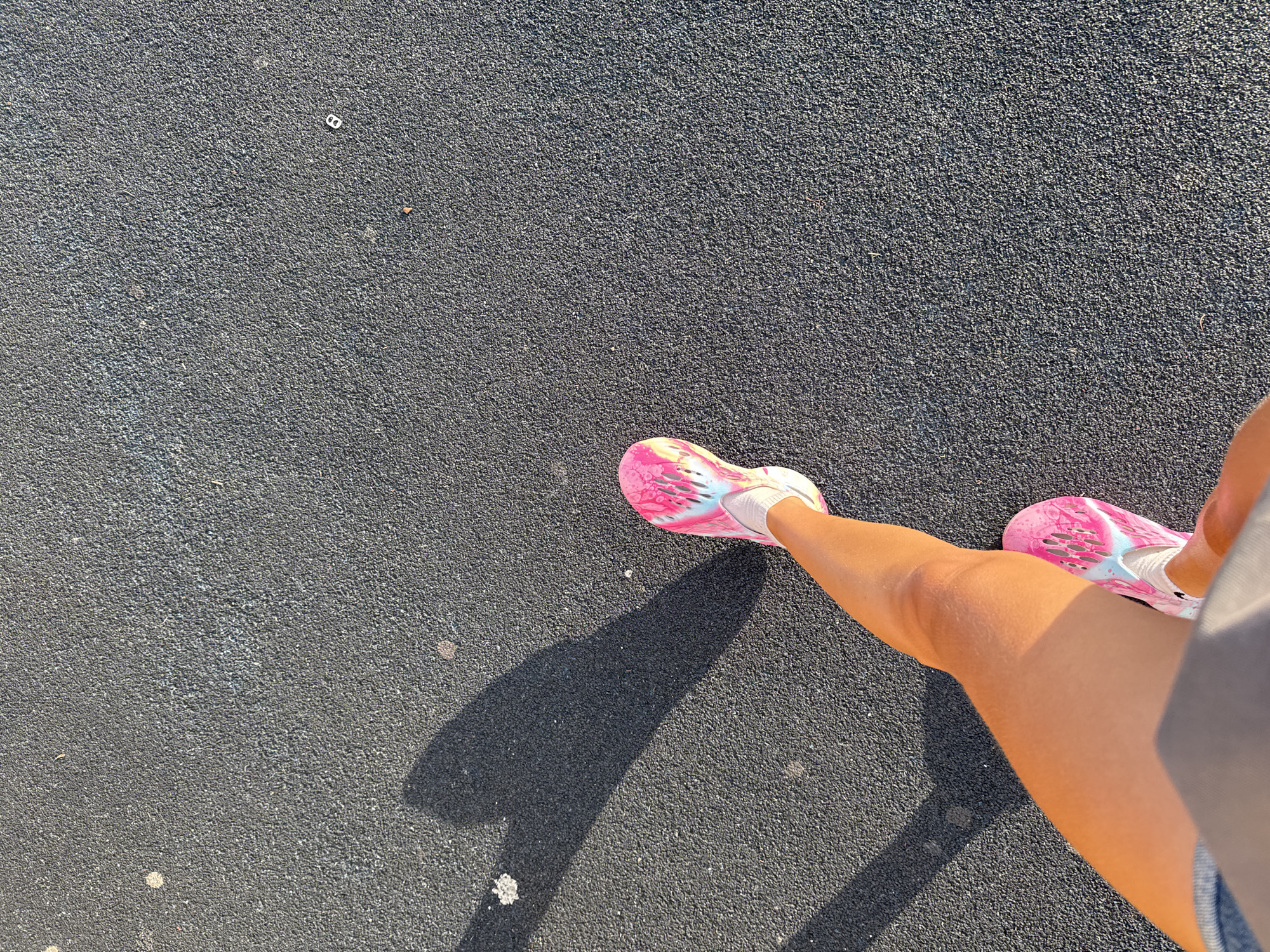 An overhead, high-angle medium shot shows a person's legs and feet walking on a dark, textured surface, likely asphalt or rubberized playground material. The person is wearing pink and white athletic shoes with a unique, abstract design. The left leg is extended forward, with the right leg positioned behind it, indicating forward motion. A shadow cast by the person on the ground resembles a heart shape, suggesting the presence of a light source behind and to the left of the subject. The environment appears to be outdoors, possibly in a park or recreational area within the city of Kurganinsk, Russia, given the context. The surface shows some small debris.
