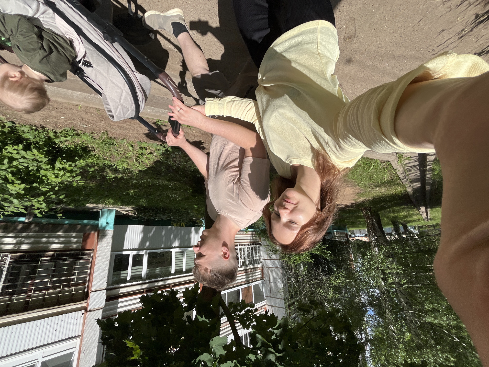 A young couple is pushing a stroller with a baby down a paved path in an urban residential area. The woman, in the foreground, is taking a selfie, smiling at the camera. She is wearing a light yellow long-sleeved shirt and black pants. The man, to her right, is looking to the side and wearing a light tan t-shirt and shorts. He is holding the stroller handles. The stroller is light gray and appears to be quilted. A baby, wearing a green shirt, is visible in the stroller. The background shows a multi-story apartment building with balconies and windows, some with metal grates. Trees and green foliage surround the path and building, suggesting a park-like setting within the city. The lighting indicates it is daytime, likely a sunny day. The scene is consistent with a residential area in Chaikovsky, Russia, characterized by apartment blocks and surrounding greenery.