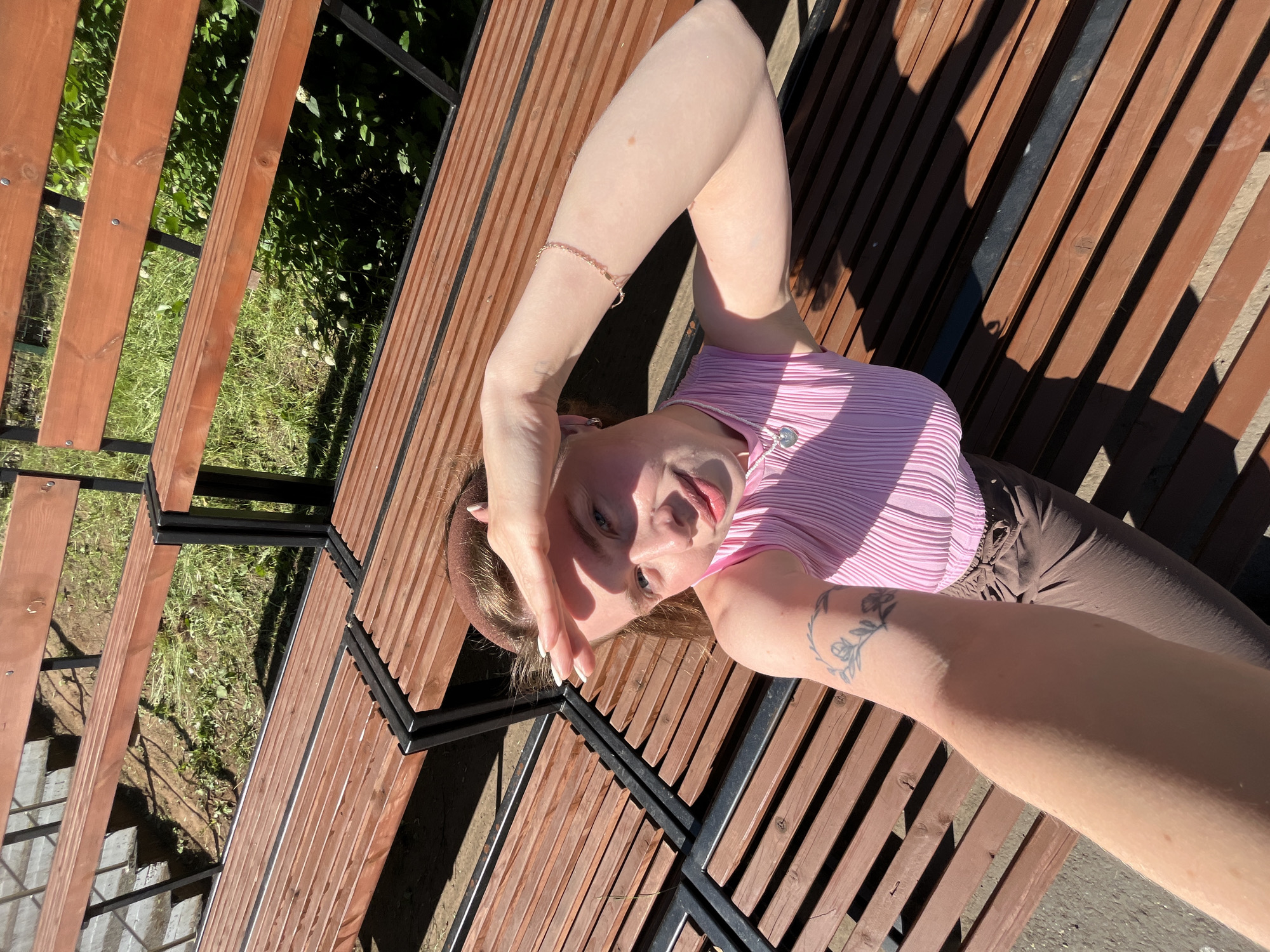 A young woman is taking a selfie while sitting on a wooden bleacher in Chaikovsky, Russia. She is wearing a pink ribbed top and brown pants. Her right hand is shielding her eyes from the sun, and a bracelet is visible on her left wrist. There is a tattoo of a circular floral design on her left forearm. The background consists of more wooden bleachers and lush green vegetation.