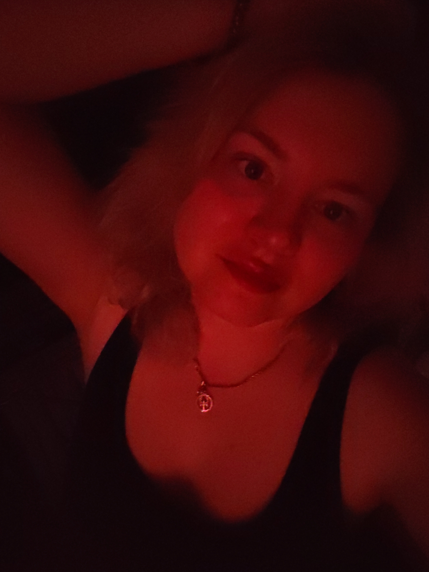 A woman with blonde hair and fair skin is posing for a selfie. She is wearing a black tank top and a gold necklace with a pendant. Her left arm is raised above her head. The photo is taken in red lighting. The location is unknown and there is no information to identify its city.