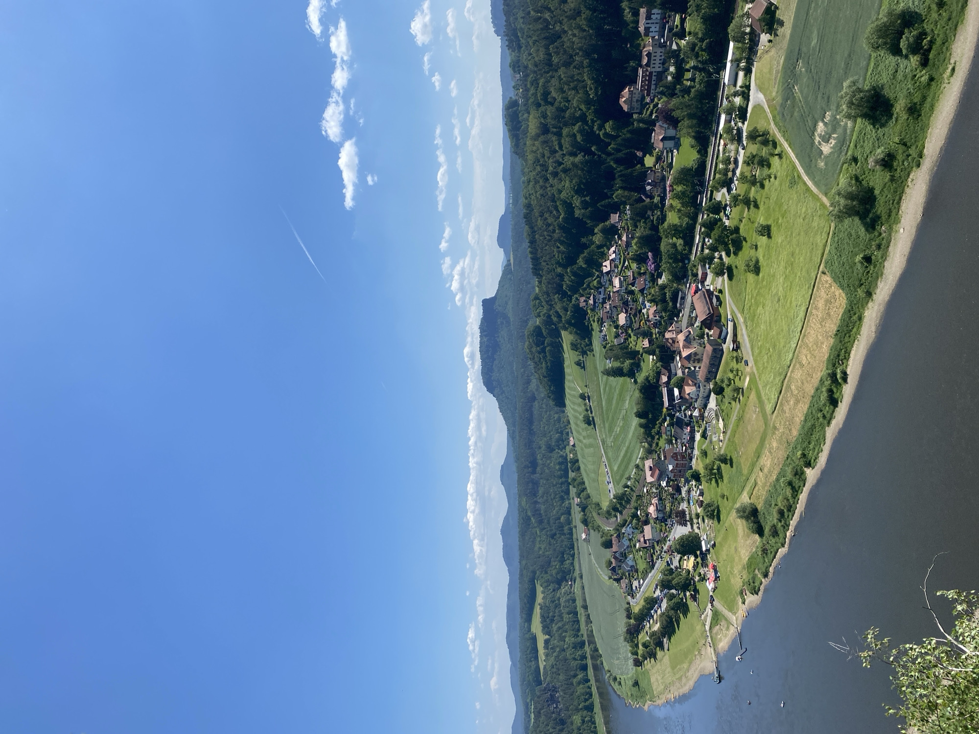 The image shows an aerial view of Lohmen, Germany, situated alongside a river. The landscape comprises a clear blue sky with scattered clouds on the left side, transitioning to a terrain of green fields, forests, and residential areas. The village architecture includes houses with red tile roofs and other buildings interspersed among the greenery. A river flows alongside the land, with a visible shoreline. The overall scene suggests a rural or small-town environment with a natural setting.