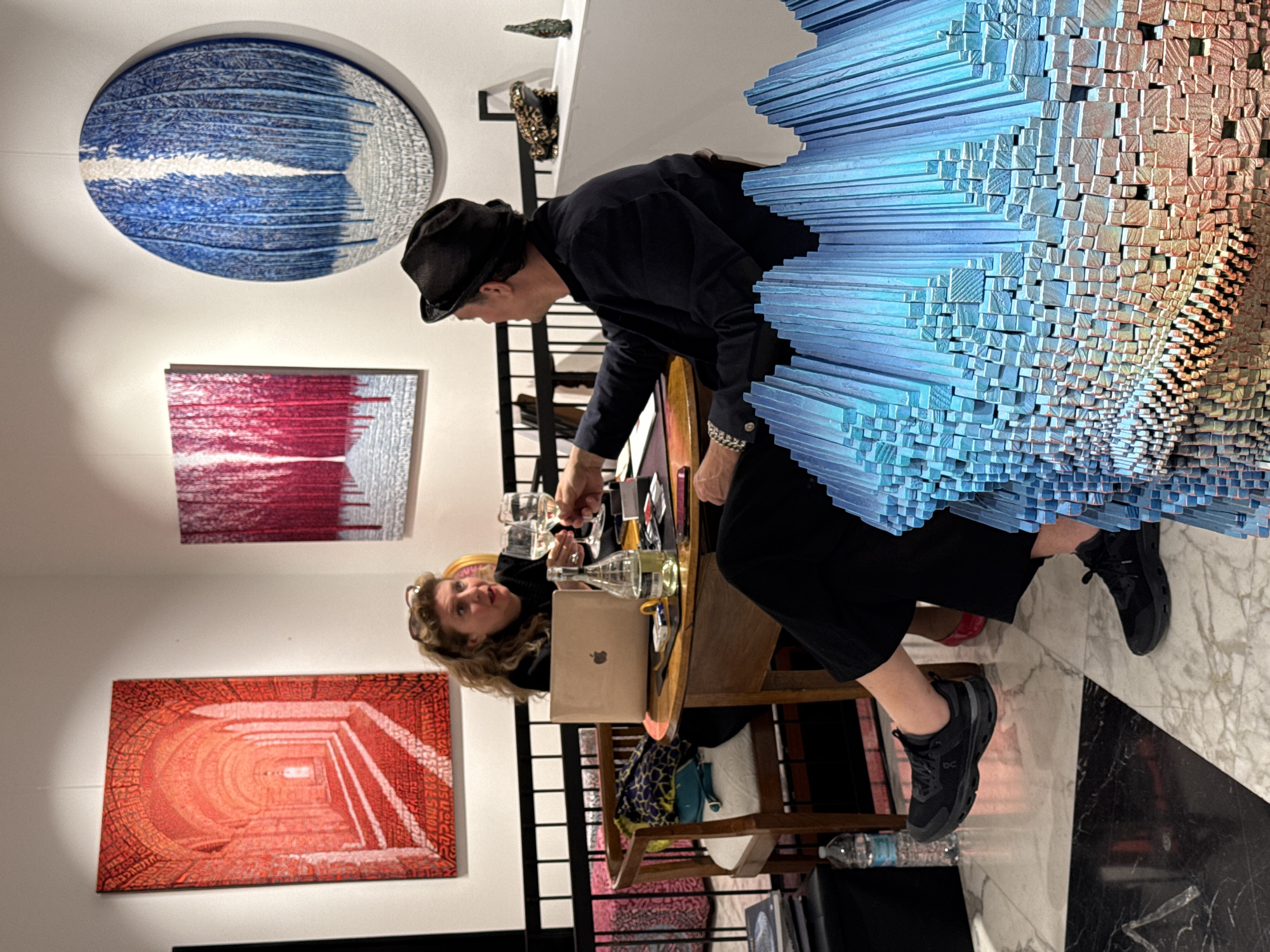 An indoor scene, likely an art gallery or exhibition in Milan, Italy, captures two individuals interacting near a wooden table, surrounded by various artworks and a large-scale sculpture.
On the left wall, three distinct artworks are displayed. The uppermost is a large circular piece, primarily blue and white, featuring horizontal textured lines. Below it, a rectangular artwork in red and white shows vertical, textured patterns. Further down, another rectangular piece depicts a vivid reddish-orange tunnel or corridor, detailed with intricate patterns or text within its depth.
In the center-left, a person wearing a dark fedora, dark jacket, and dark trousers is leaning forward over a wooden table, appearing focused on something. A patterned bracelet is visible on their left wrist. Seated at the table is another person, a woman with long, curly blonde hair, dressed in a dark top. She holds a glass and looks up and slightly to the right, observing or conversing. The table holds wine glasses, bottles (one with white wine), and a silver laptop bearing the Apple logo.
Dominating the right side of the frame is a large, abstract sculpture composed of numerous small, rectangular blocks or planks. These blocks are arranged in a dynamic, swirling, layered pattern, transitioning in color from cool blue to warmer, reddish-brown tones towards the center.
The flooring is a patterned black and white marble. Both individuals wear black sneakers; the woman's shoe clearly shows the "ON" brand logo. A plastic water bottle rests on the floor beside the table. The overall lighting suggests an indoor setting, possibly an evening event.